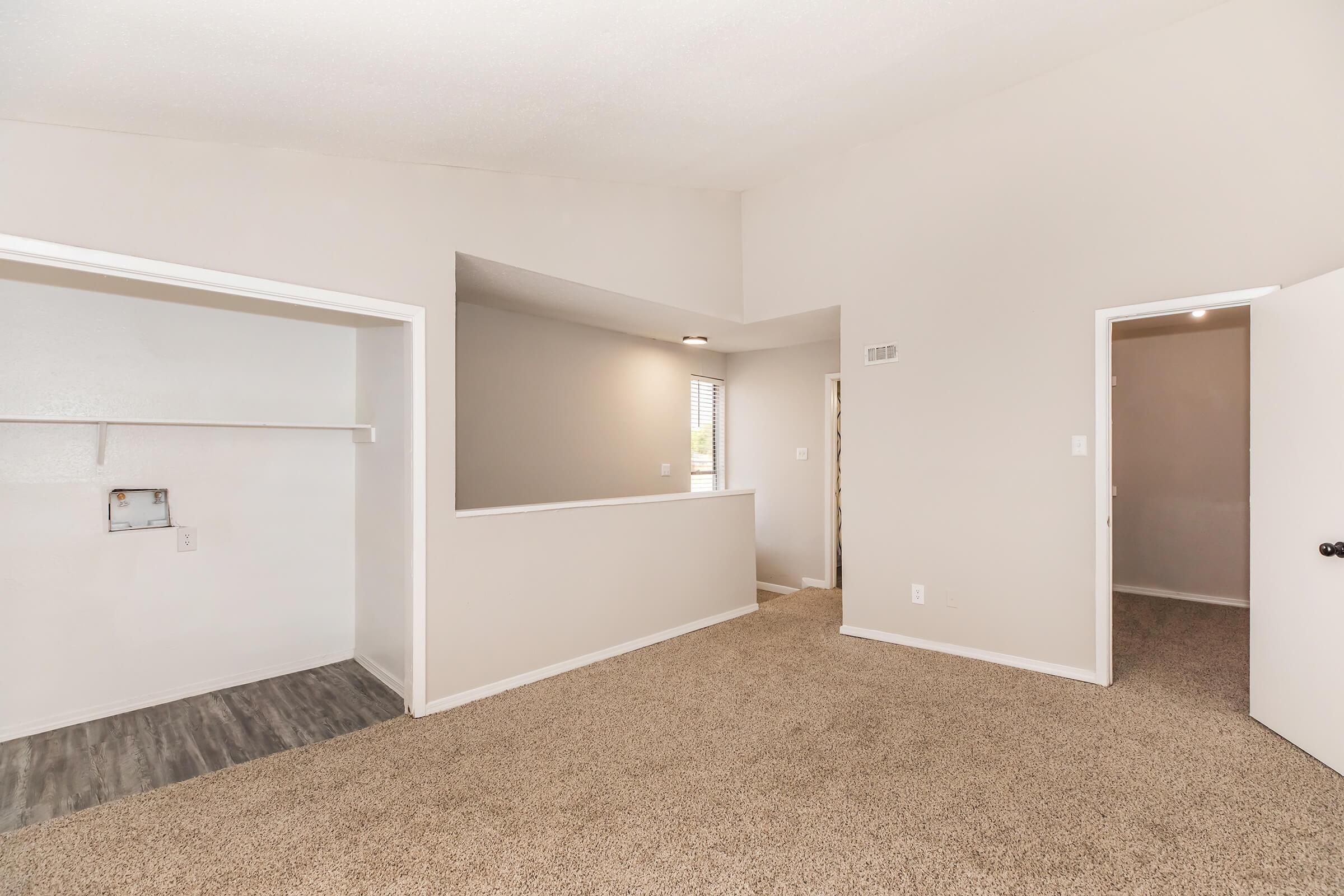 Interior view of a spacious living area with beige carpet and light-colored walls. To the left, there is a walk-in closet; in the center, a half-wall creates an open feel. A doorway on the right leads to another room. The space is well-lit with natural light, creating a warm and inviting atmosphere.