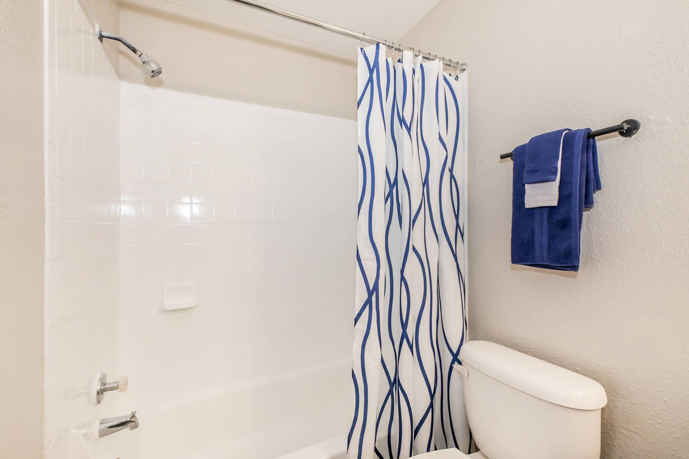 A clean bathroom featuring a shower with a white curtain adorned with blue wavy patterns, a toilet, and a towel rack holding a folded blue towel. The walls are a neutral color, and the overall atmosphere is tidy and inviting.