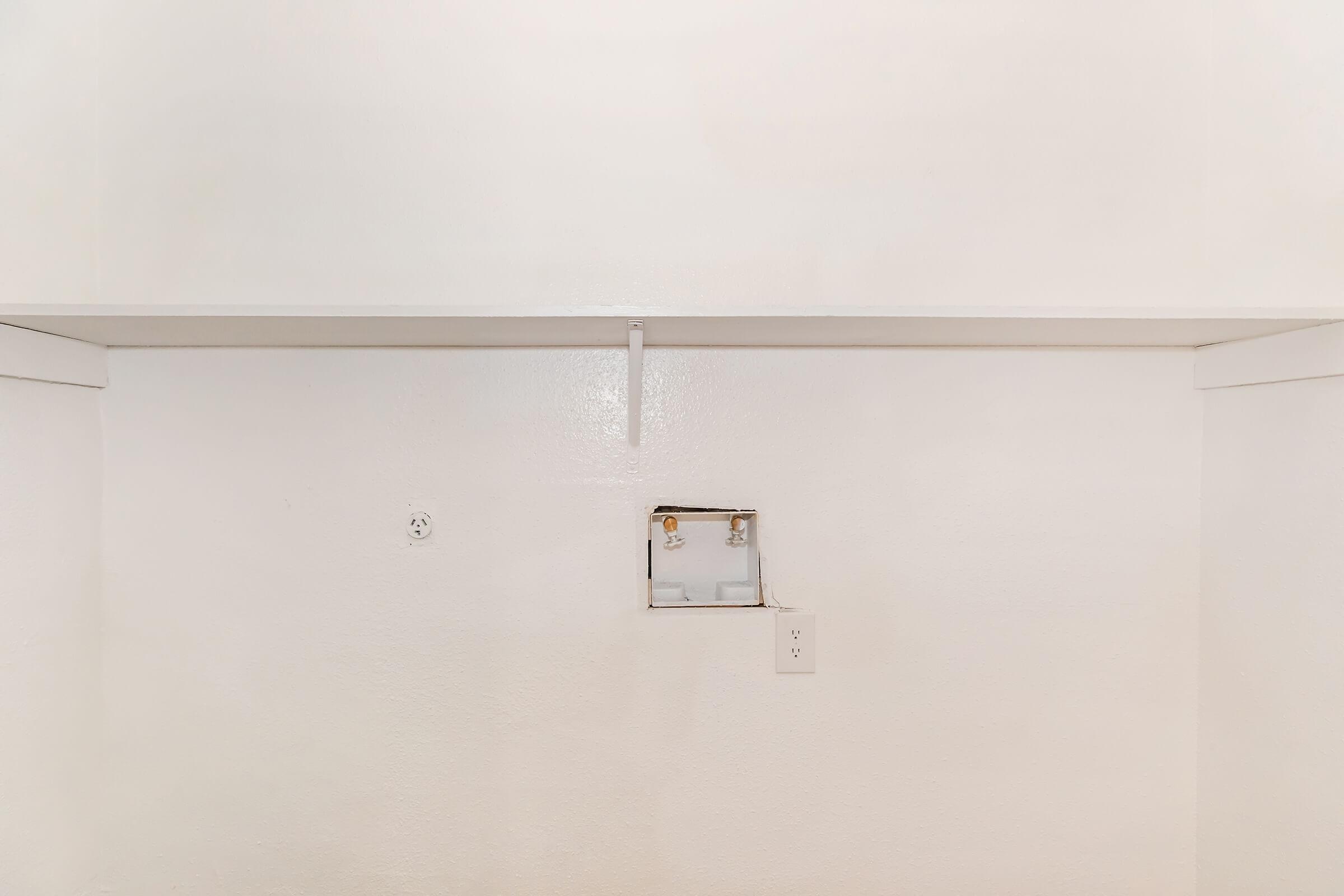 A minimalistic white wall with a small patch indicating a removed fixture. There is a shelf above the patch, and a single electrical outlet is visible. The overall space appears empty and clean, suggesting a simple room or closet without decorations.