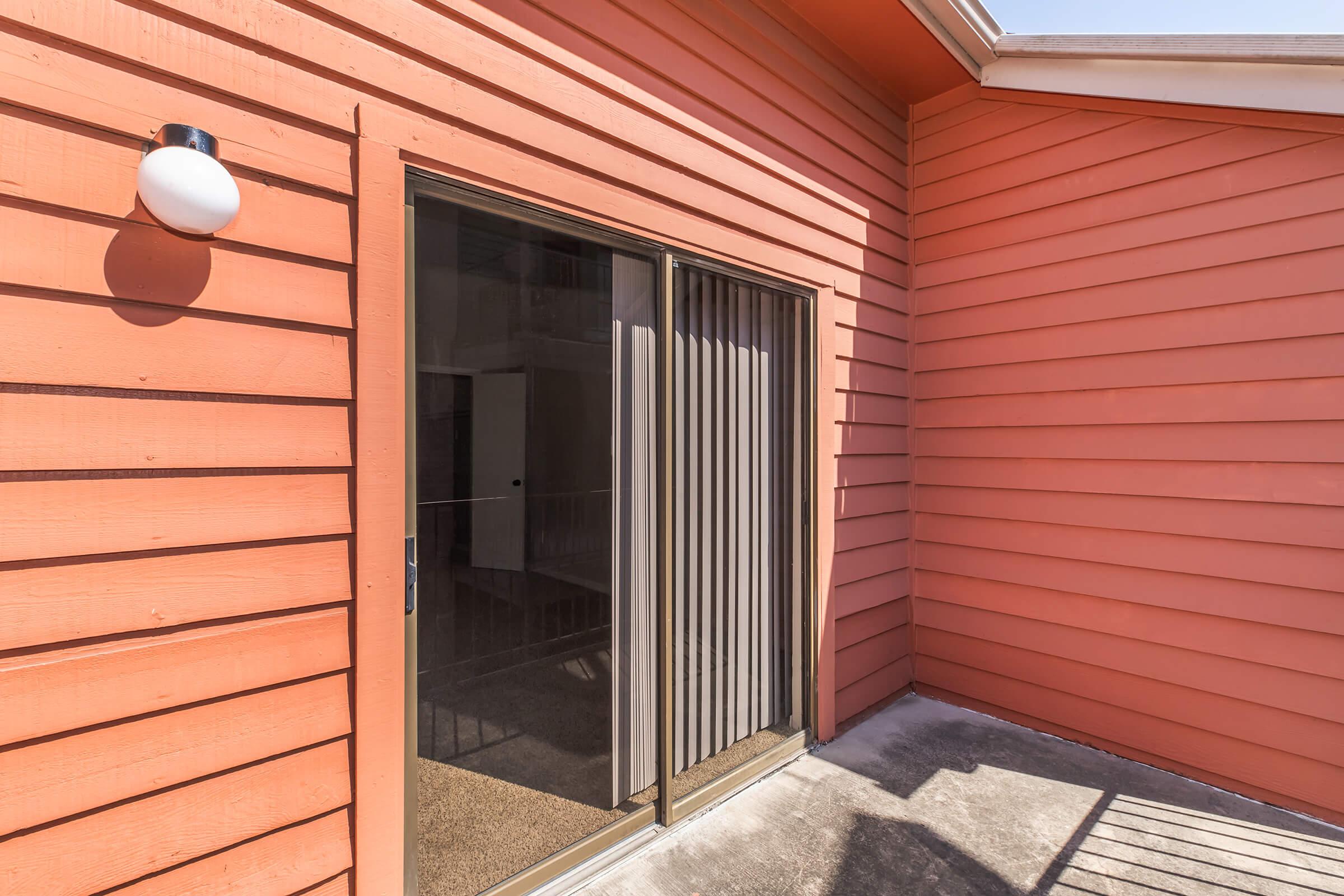 A close-up view of a sliding glass door with vertical blinds, leading to an interior space. The door is framed by orange wooden siding, and a wall-mounted light fixture is visible on the left. The setting appears to be a patio or balcony area, with a concrete floor.