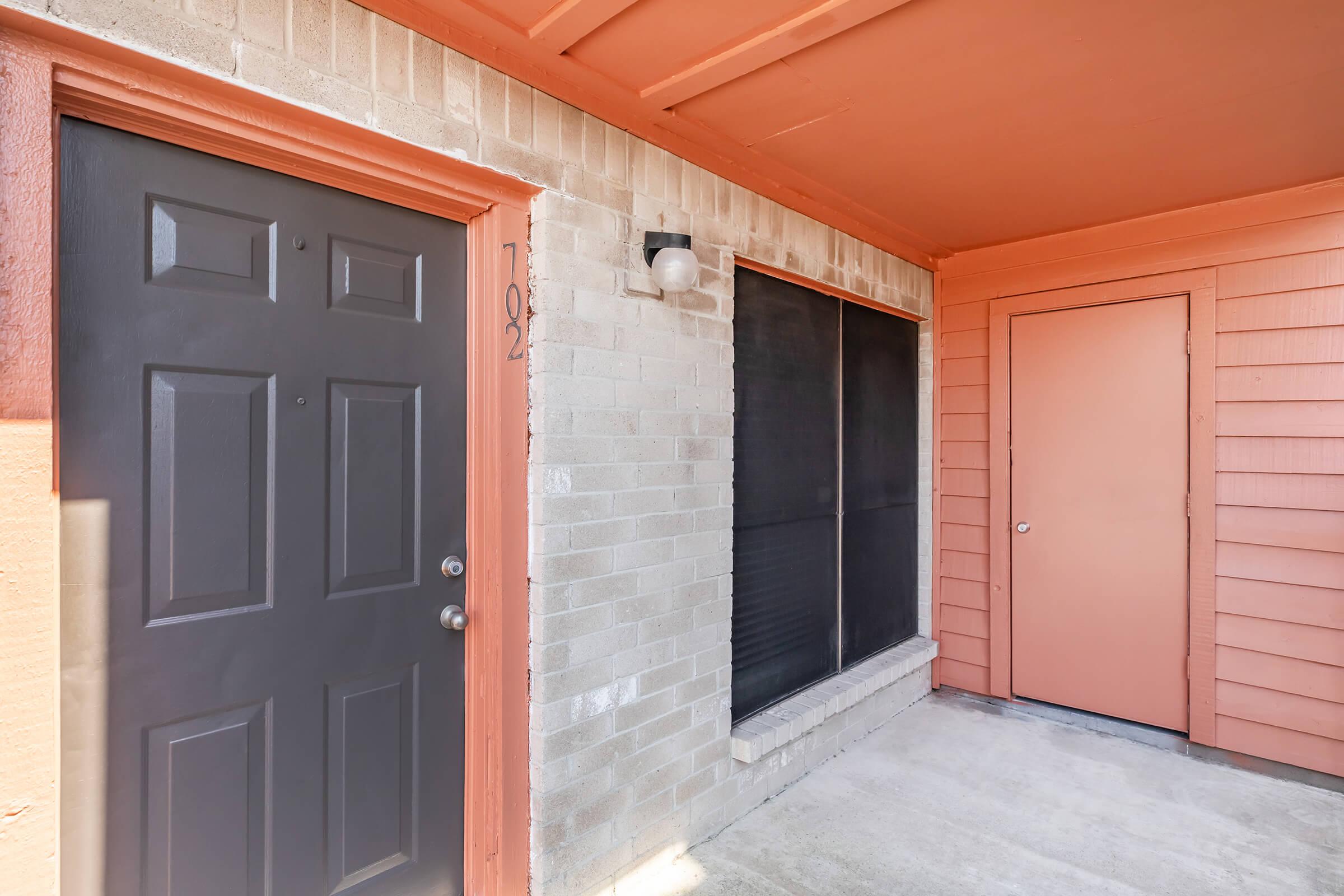 A view of an apartment entrance featuring a dark gray door labeled "102," a light brick wall, and an adjacent window covered with a black screen. The wall is accented with orange trim, and there is an additional orange door visible nearby, creating a vibrant and inviting entrance area.