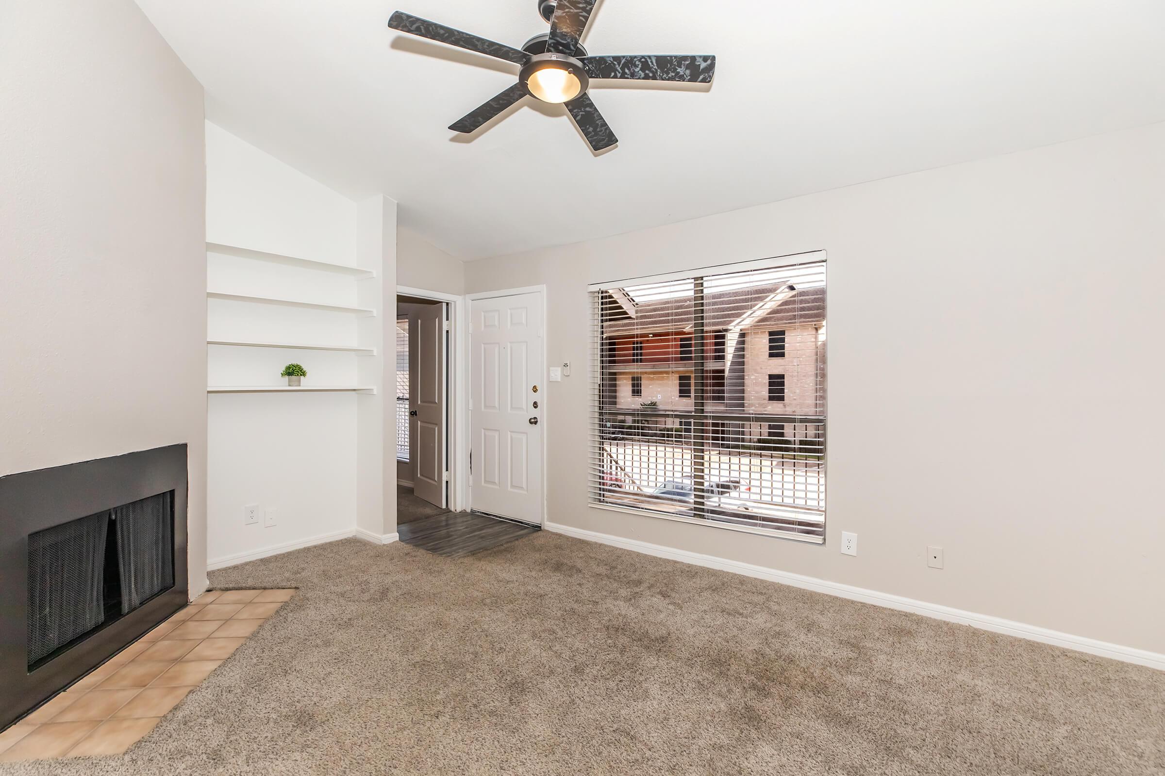 A cozy living room featuring light beige walls, plush carpet, and a ceiling fan. There’s a decorative fireplace with a tiled base, built-in shelves on one side, and a large window with horizontal blinds offering a view of the outside. A door leads to the entrance, enhancing the space's inviting atmosphere.