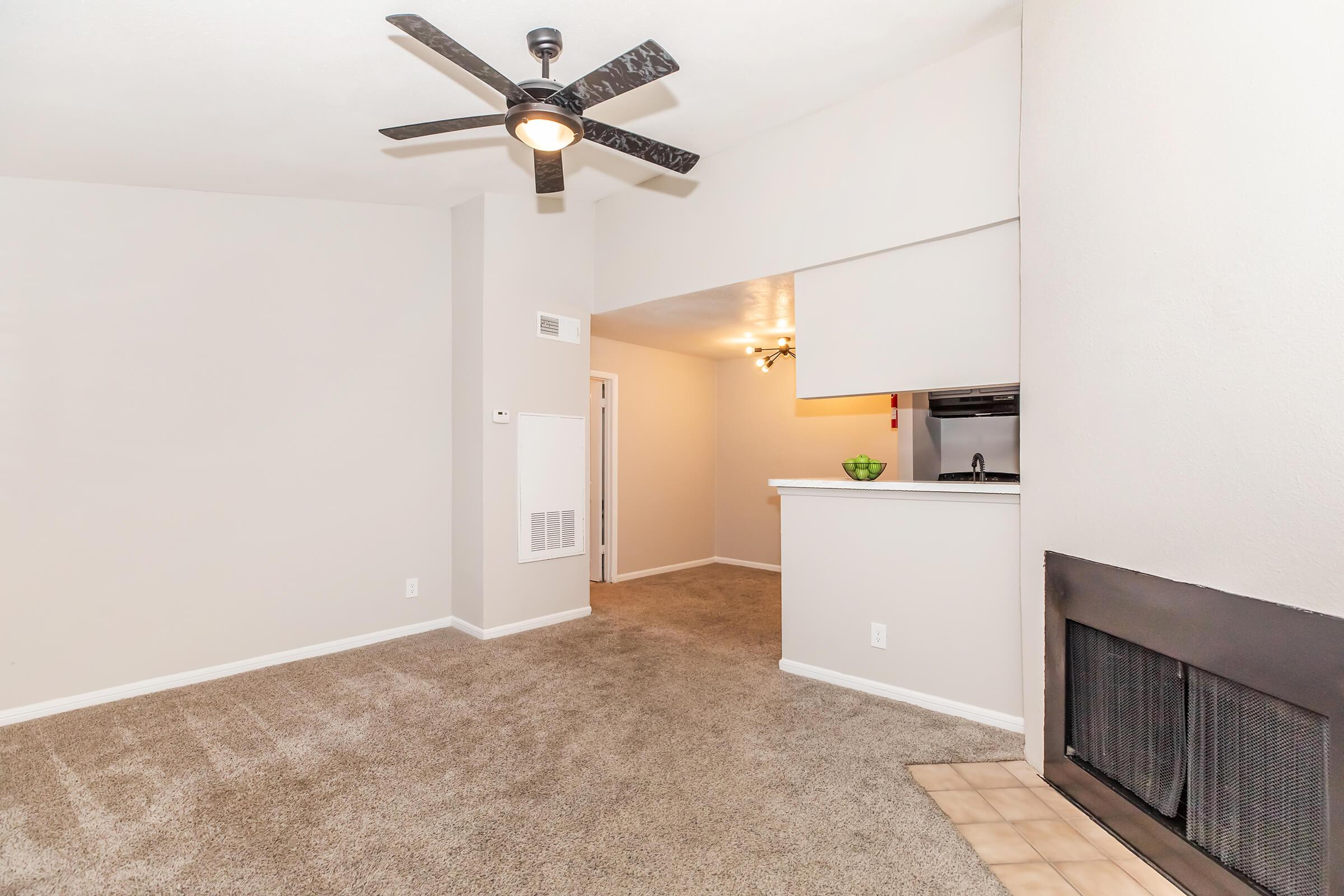 A spacious, well-lit living room featuring light gray walls, plush carpet, and a ceiling fan. A small kitchenette is visible in the background, with a bar-height counter separating it from the living area. A fireplace is located on the right, adding warmth to the space.