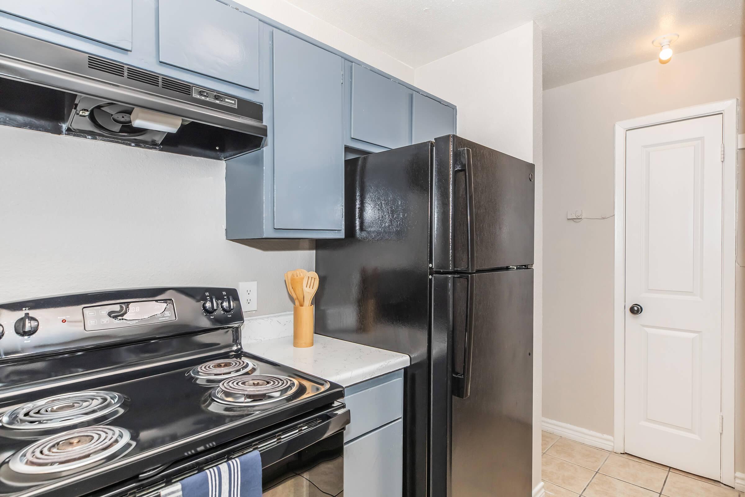 A modern kitchen featuring a black stove with four burners, a black refrigerator, and blue cabinetry. Countertops are light-colored, and cooking utensils are displayed in a wooden holder. A white door leads to another room, and the floor is tiled in beige. The overall space is well-lit and sleek.