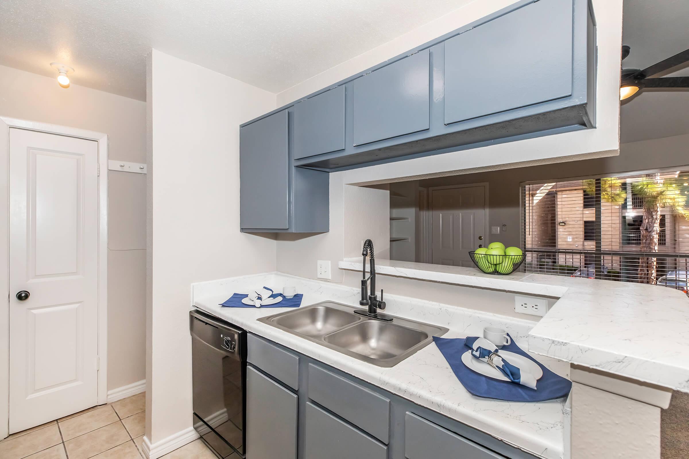 A modern kitchen featuring gray cabinets, a double sink with a black faucet, and a silver dishwasher. There is a small window with a view of an outdoor area, and two blue napkins are placed beside the sink on a white countertop. The walls are painted in a light color, creating a bright, airy atmosphere.