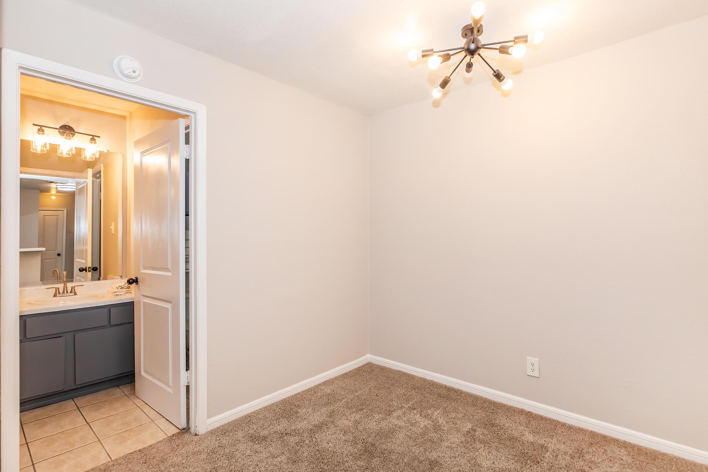 A well-lit room featuring beige walls and plush carpet. A doorway leads to a bathroom with vanity and mirror. A modern chandelier hangs from the ceiling, casting soft light. The overall atmosphere is clean and inviting, suitable for a bedroom or living space.