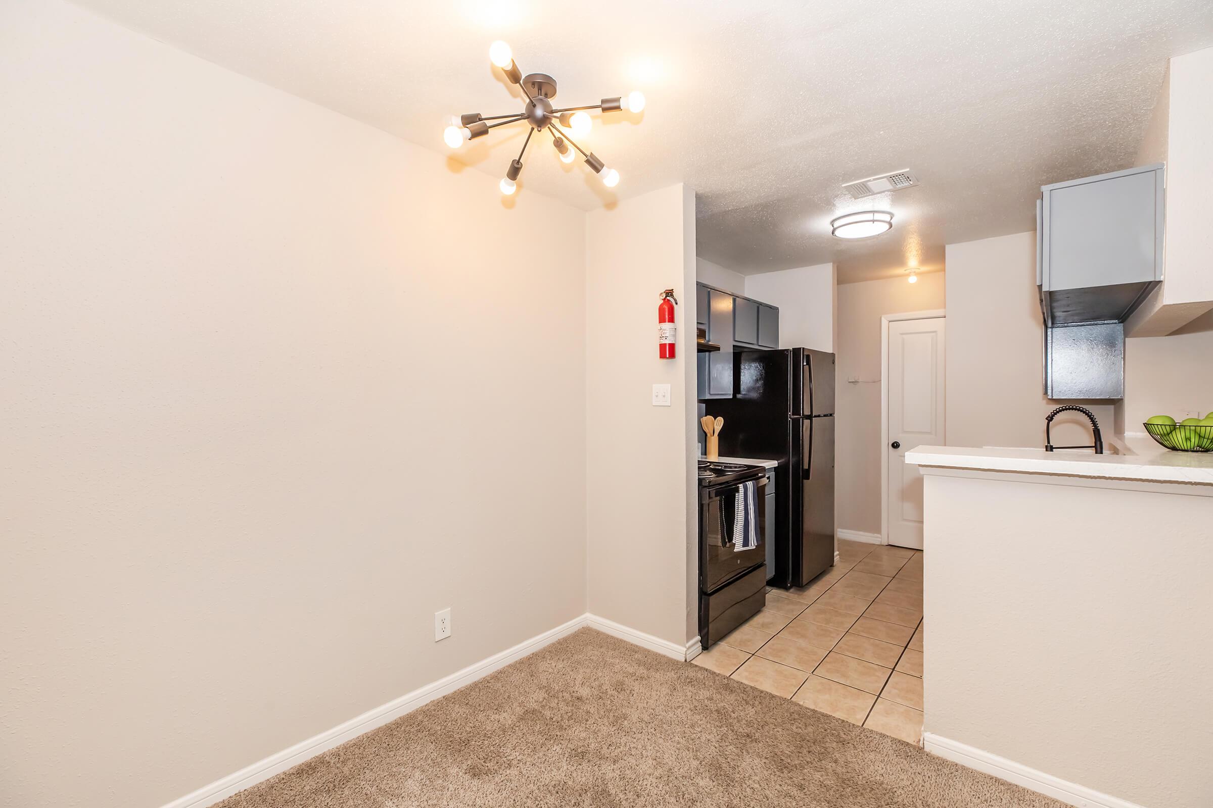 Interior view of a modern kitchen and dining area featuring beige walls, a contemporary light fixture, and a tan carpet. The kitchen includes dark appliances, white cabinets, and a countertop. The space appears bright and inviting, with a doorway leading to another room visible in the background.