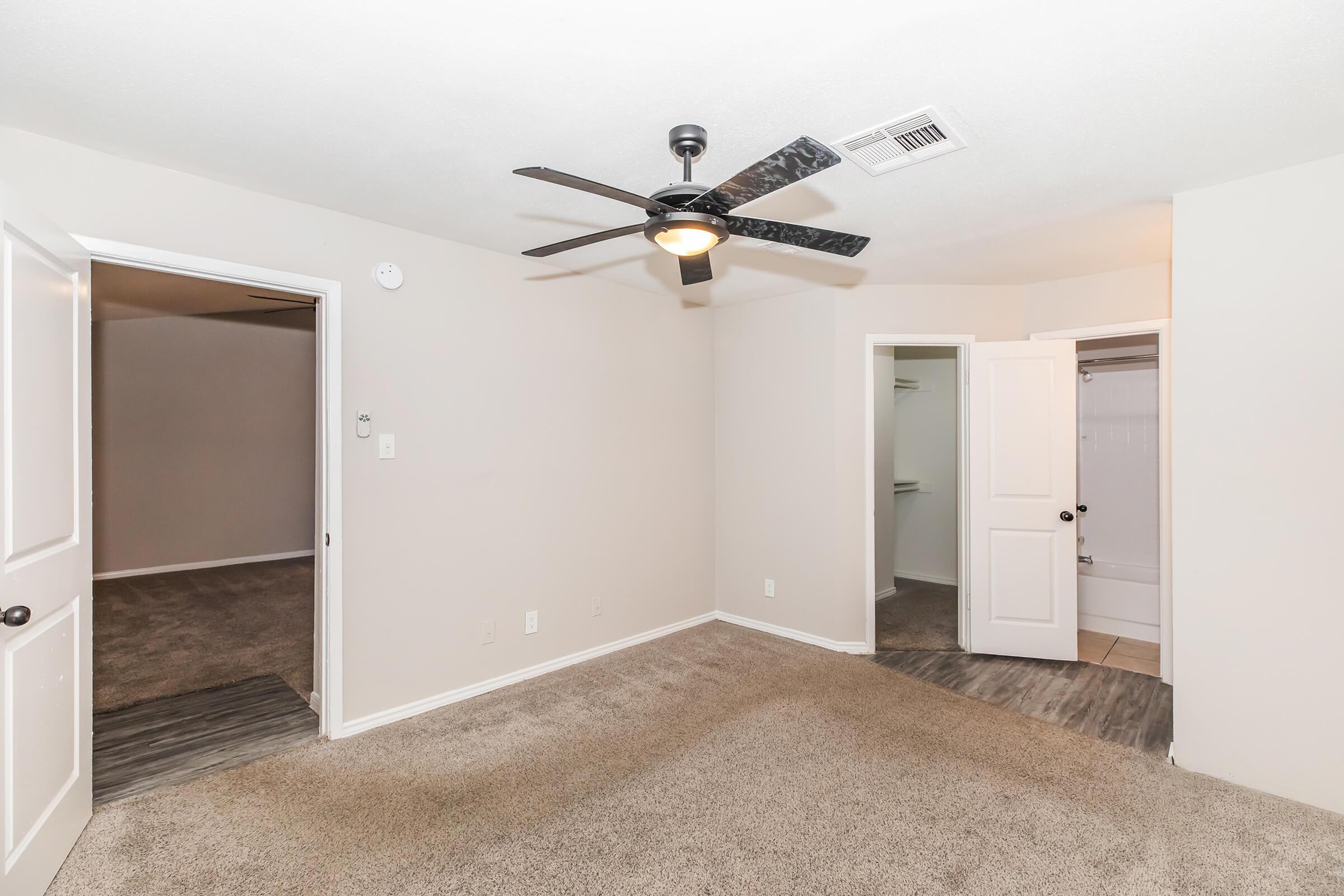 A neutral-toned room featuring a ceiling fan, carpets, and doors leading to other spaces. On the left, there is an open doorway leading to another room, and on the right, a door opens to a bathroom area. The walls are painted light shades, creating an airy atmosphere.
