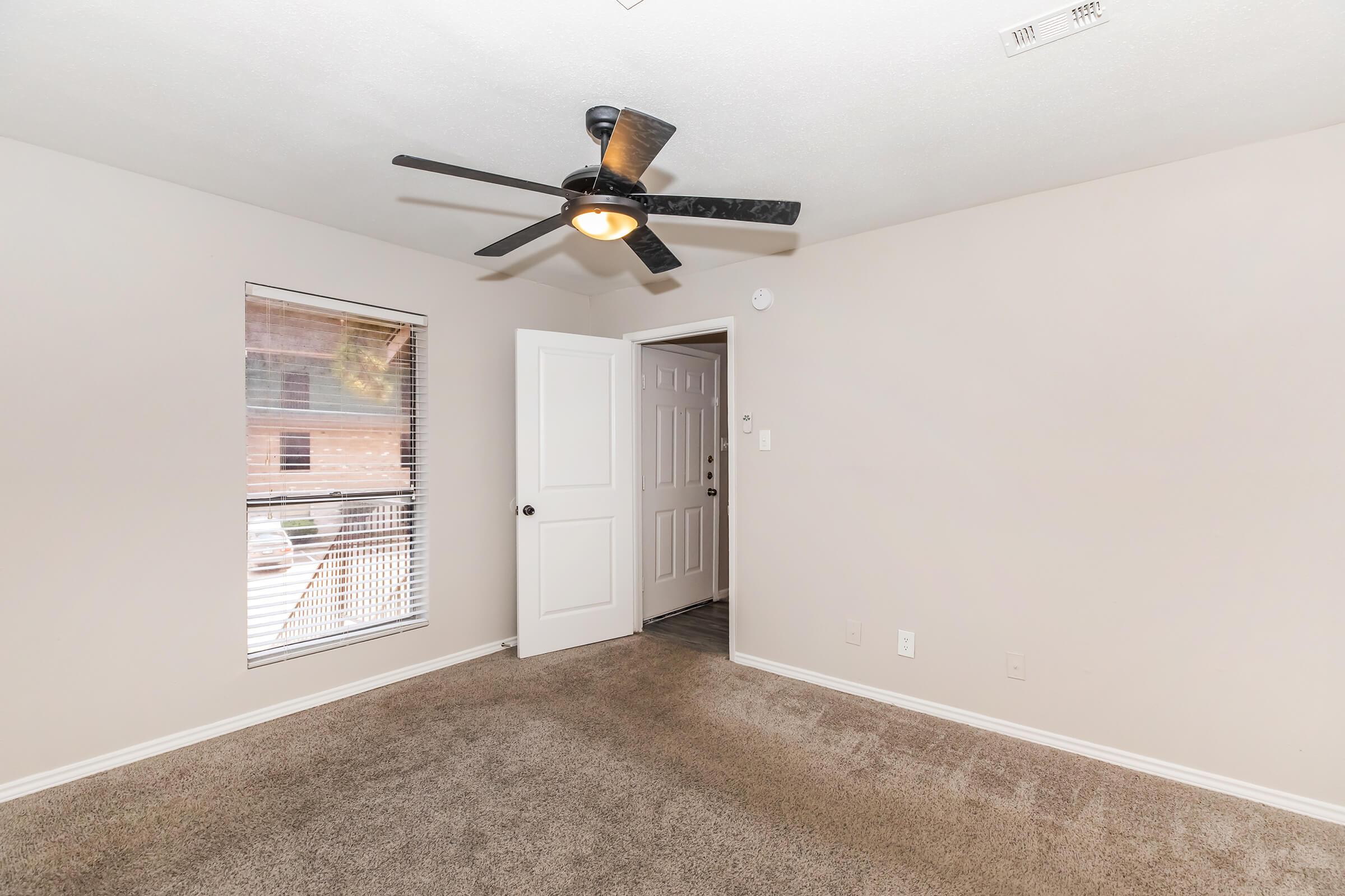 A well-lit, neutral-colored room with carpeted flooring, featuring a ceiling fan and a window with blinds. The door prominently leads to another area, enhancing the spacious feel of the room. The walls are a light shade, creating an inviting atmosphere.