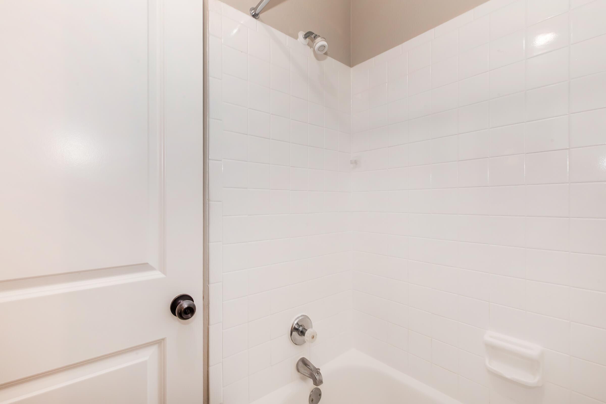 A clean, modern bathroom featuring a white tiled shower area with a showerhead, a chrome faucet, and a flat soap dish. The door to the bathroom is partially visible, showing a white panel door with a round doorknob. The overall aesthetic is bright and minimalist.