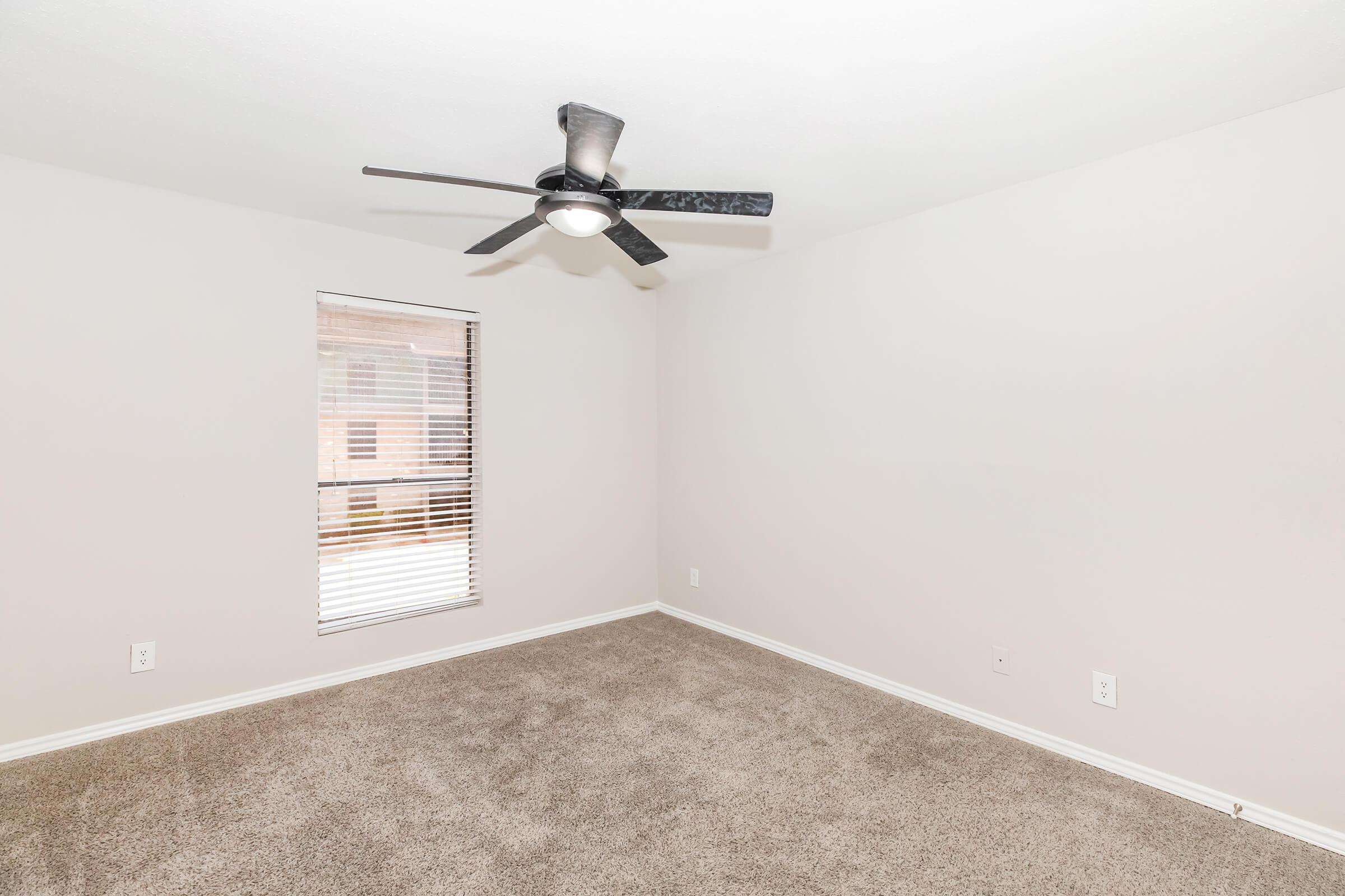 A vacant room with light-colored walls and carpet. There is a ceiling fan with dark blades, and a window with blinds allowing natural light to enter. The space is tidy and features neutral decor, creating a bright and open atmosphere.