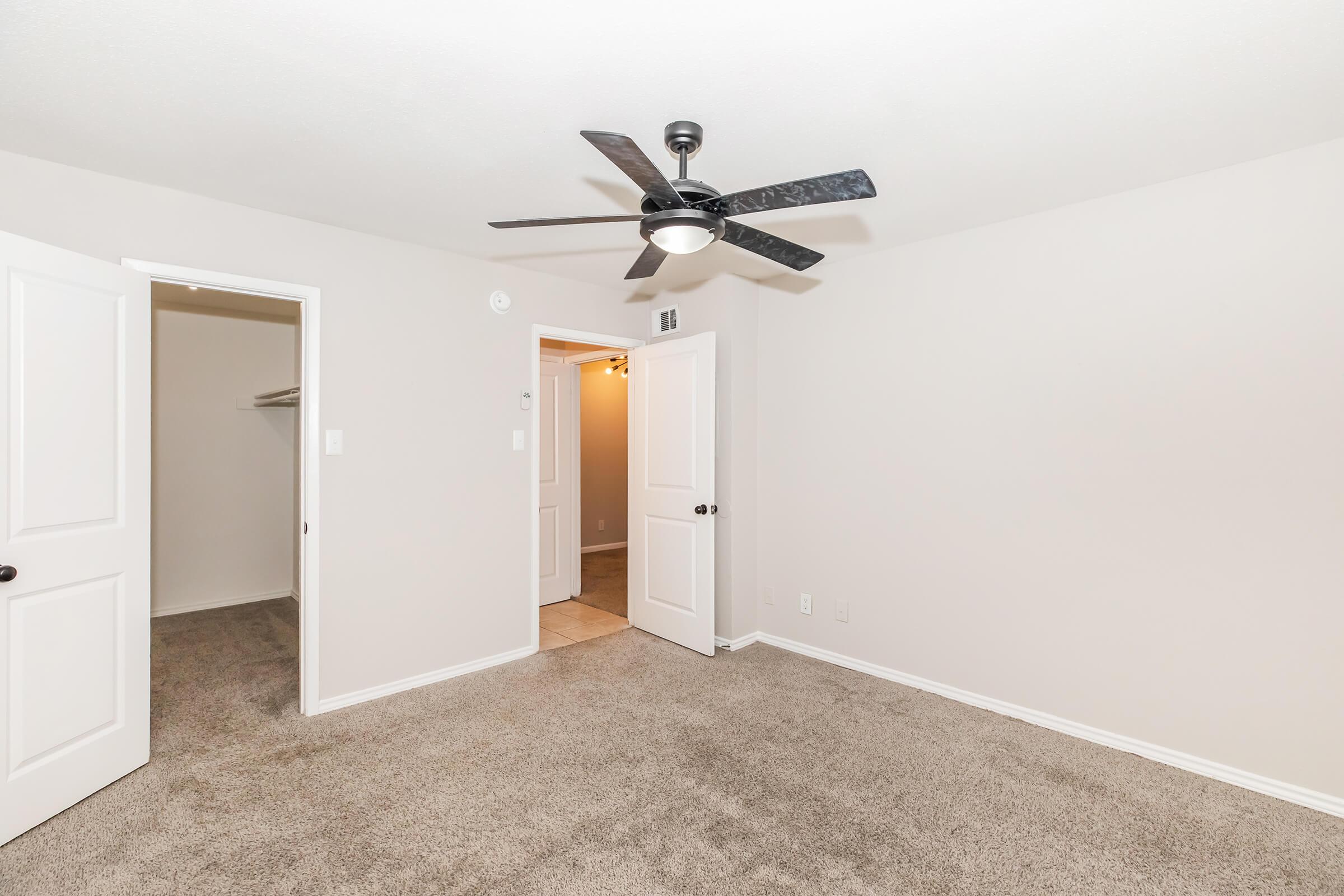 A spacious room featuring a light-colored carpet, a ceiling fan with dark blades, and white doors leading to a closet and an adjoining room. The walls are painted in a neutral tone, creating a bright and airy atmosphere. The room is well-lit with natural light coming from the partially visible entrance.