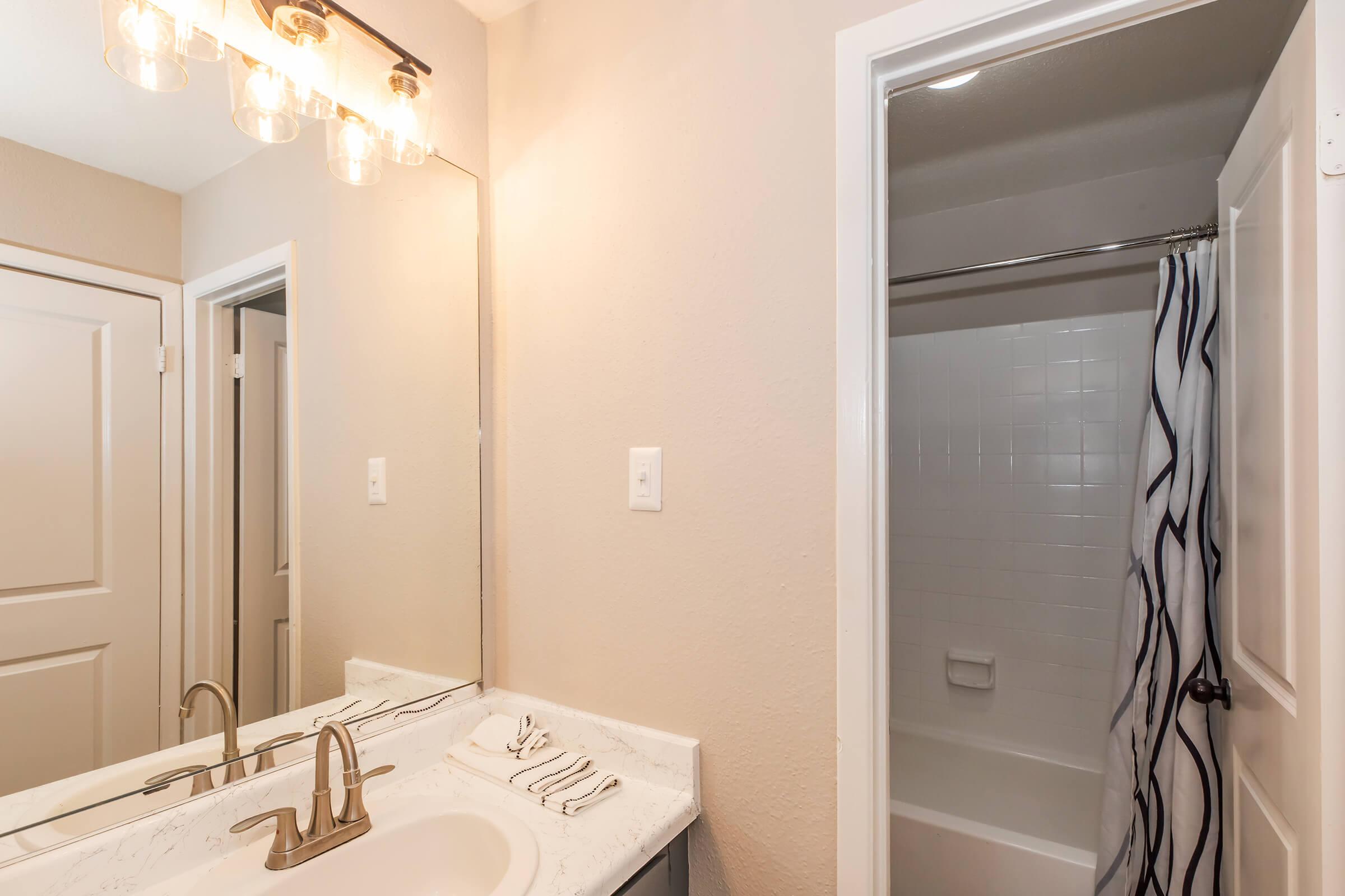 A well-lit bathroom featuring a vanity with a sink and modern faucet, a large mirror above the sink, and a shower area with a curtain. The walls are painted a light color, and the flooring appears to be a clean tile. Two doors are visible, leading to separate areas.