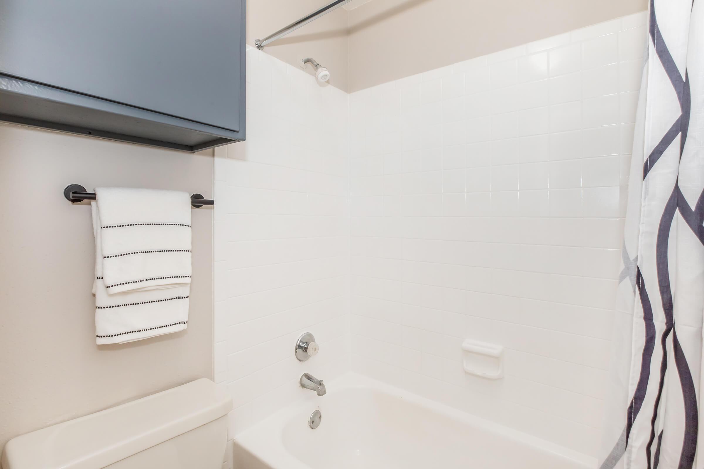Clean and modern bathroom featuring a white tiled bathtub and shower, with a sleek silver faucet. A black cabinet is mounted above, and a towel bar holds neatly folded white towels with black stripes. The shower curtain is also white, adding to the minimalist aesthetic of the space.