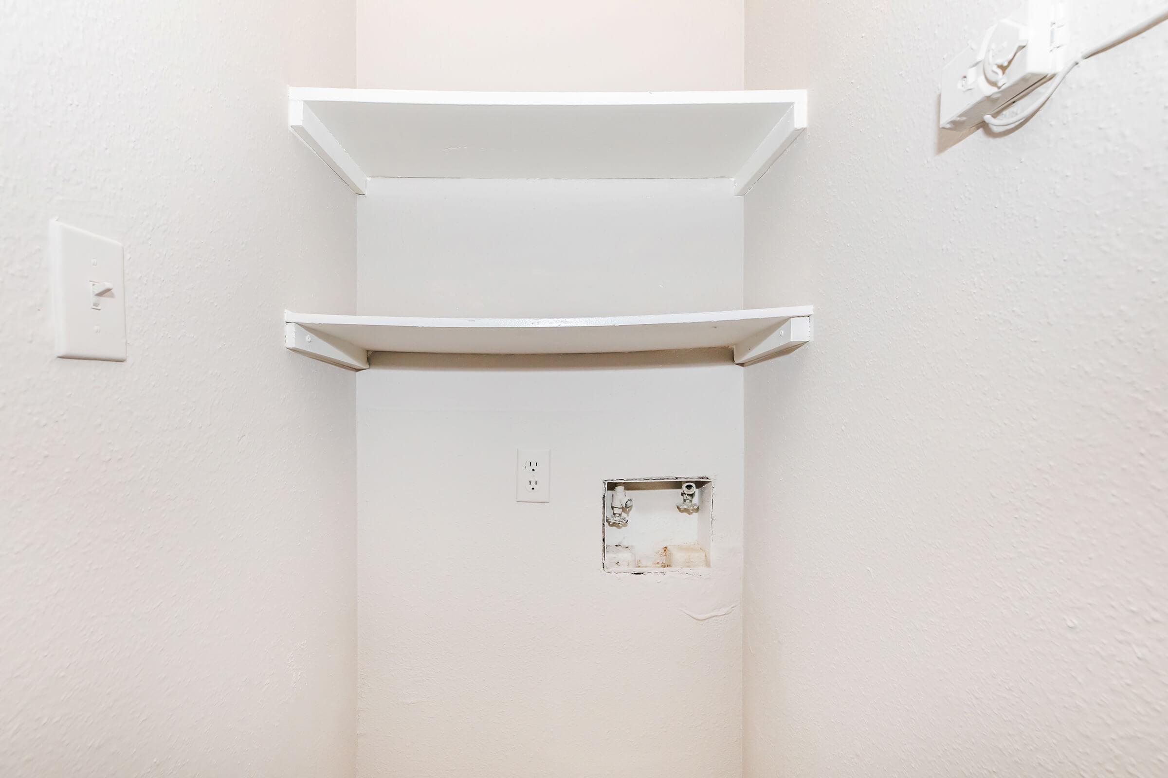 A small, empty closet with white walls and shelves. The shelves are positioned at different heights, and there is a small opening in the wall with visible electrical outlets. The floor is bare and there are no additional items or decorations present.