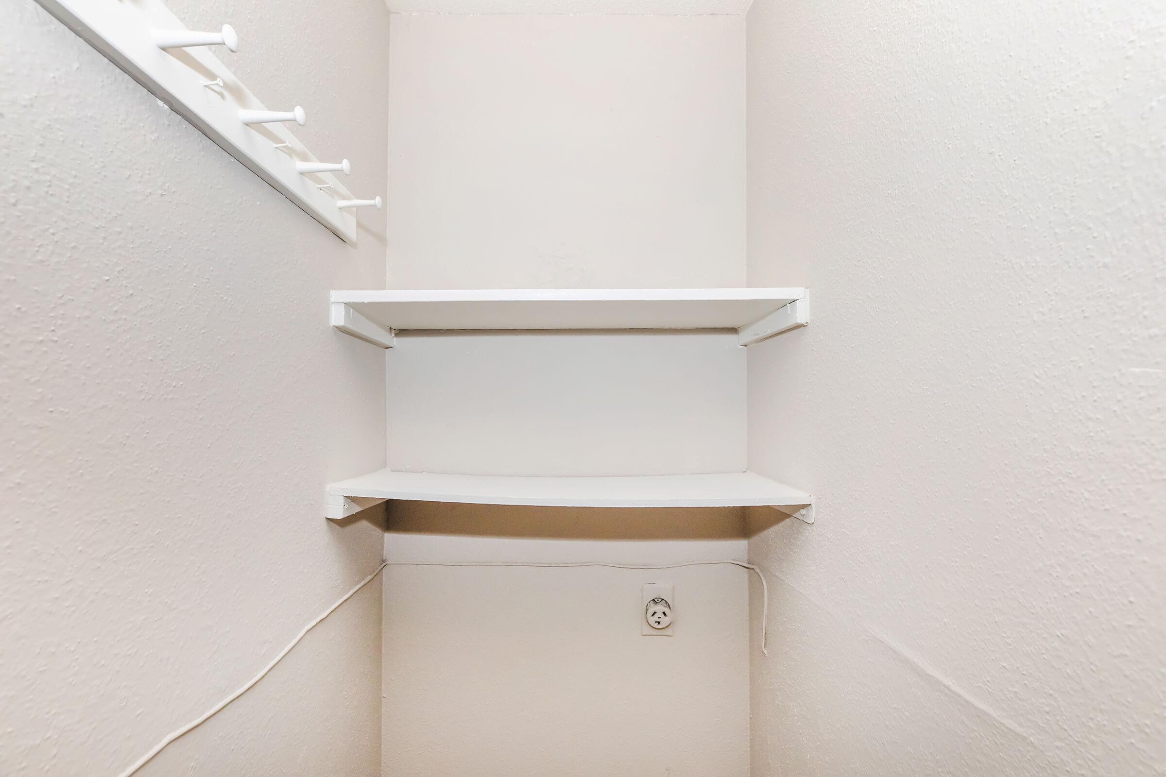 A small, empty storage space with two white shelves against the wall. There are hooks mounted above the shelves, and a power outlet is visible near the floor. The walls are plain and painted in a light color, creating a clean and minimalist appearance.