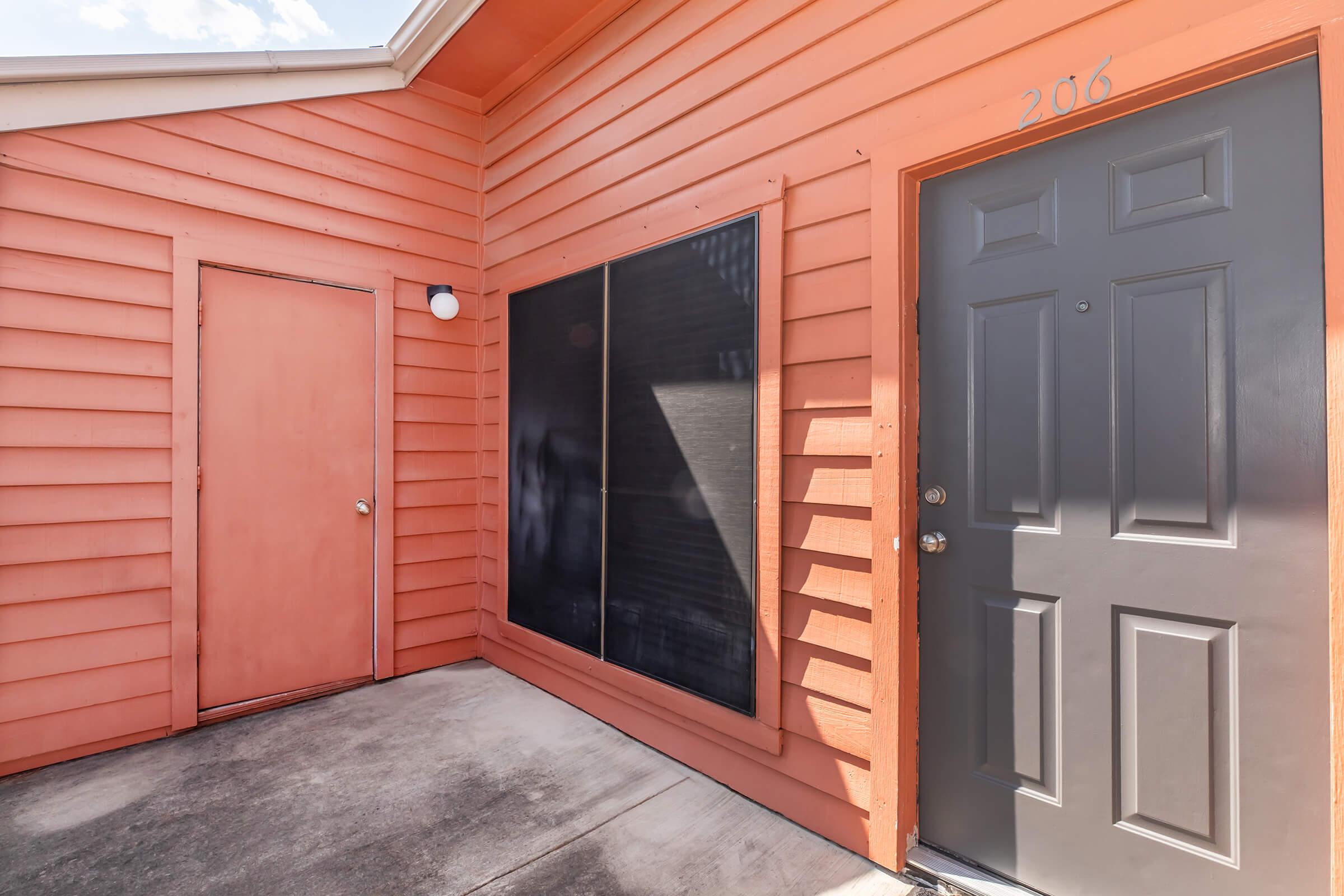 A bright orange exterior of a building with two doors—a dark grey door labeled "206" and a smaller pink door to the left. There is a large window covered by a black screen, and a light fixture above the grey door. The ground is concrete, and the sky is visible in the background.
