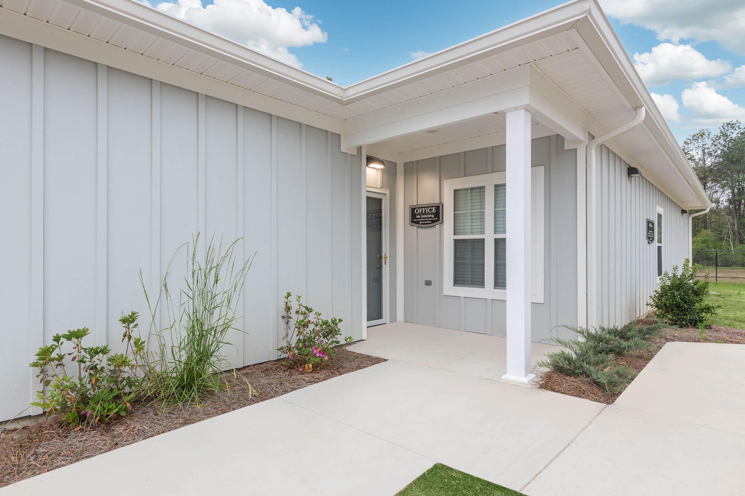 A modern office building featuring a light gray exterior with a welcoming front entrance. The porch has two large windows and a sign labeled "Office." The landscape includes some light greenery and a clean concrete pathway leading to the entrance, under a bright blue sky with fluffy white clouds.