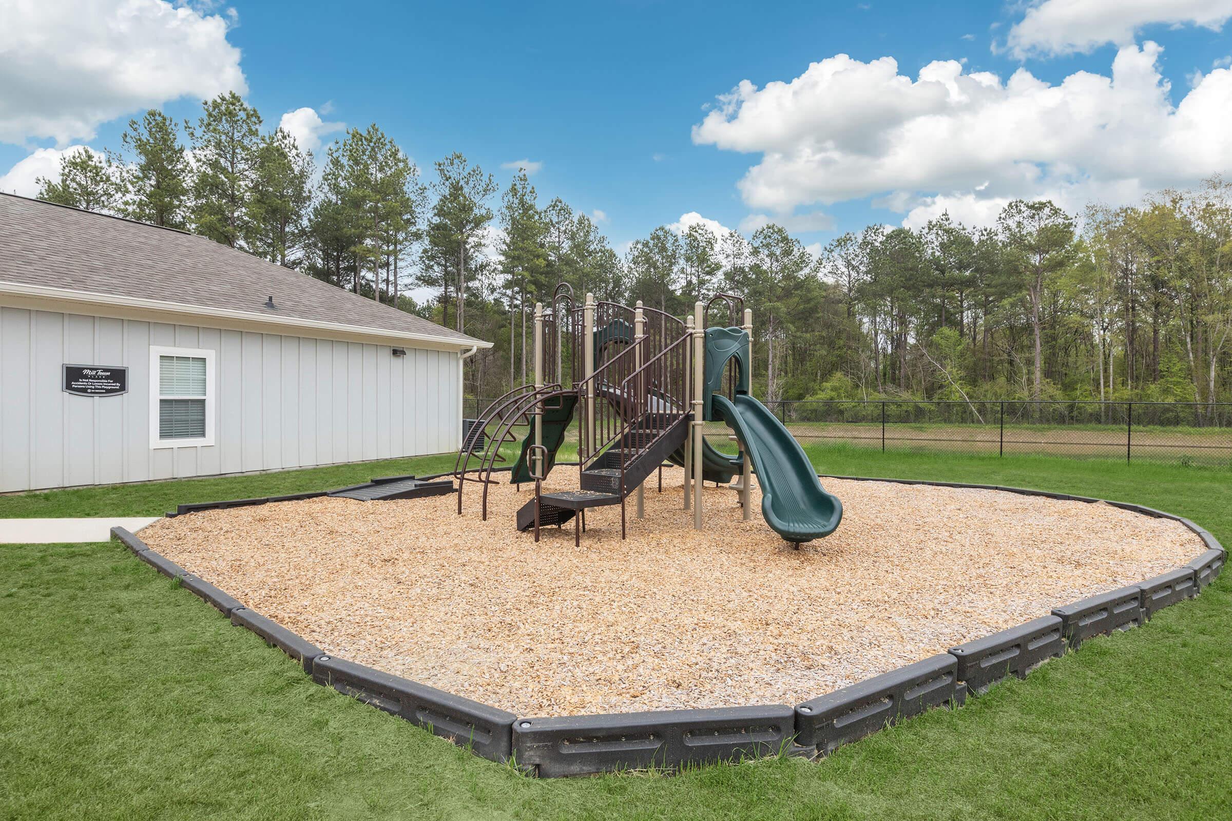 Playground area featuring a combination of slides, climbing structures, and swings, surrounded by a soft wood chip ground cover. The playground is located beside a white building, with green grass and trees in the background under a partly cloudy sky. Safe and inviting for children.