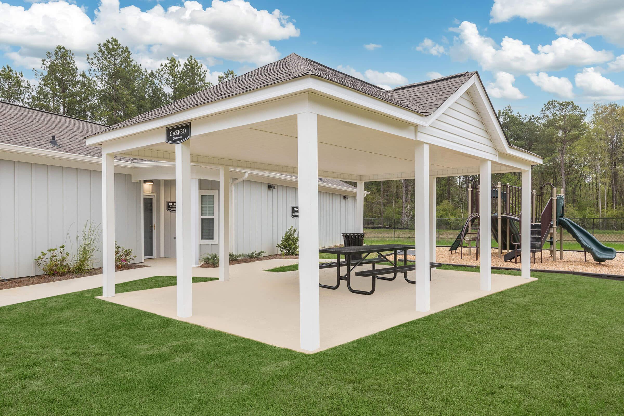 A shaded picnic area featuring a white gazebo with a picnic table, surrounded by green grass. In the background, there are two playground structures, including a slide, and a building with multiple entrance doors under a blue sky with scattered clouds.