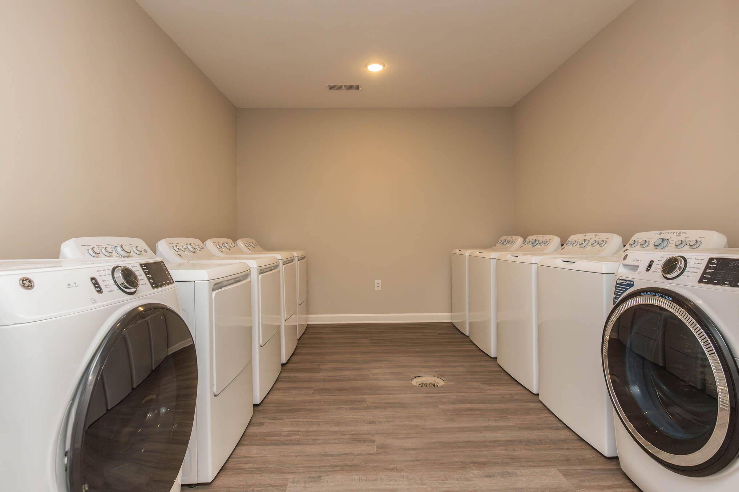 A spacious laundry room featuring multiple white washing machines and dryers arranged in a row on a wooden floor. The walls are painted in a light neutral color, and the room is well-lit with ceiling lights. There are no windows visible in the image.