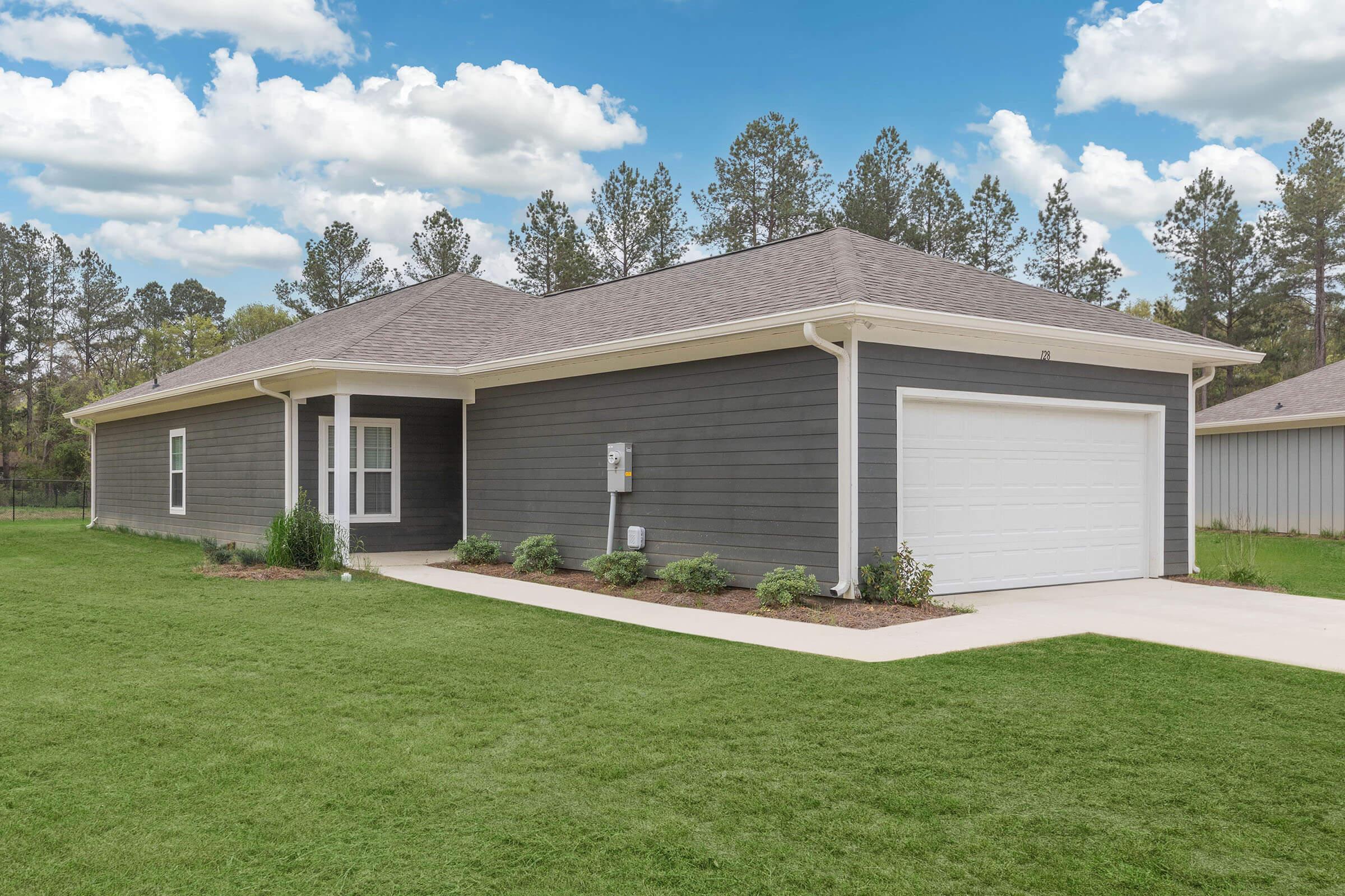 A modern, single-story house with gray siding and a white garage door, surrounded by a green lawn. The home features a covered porch area and a pathway leading to the entrance, with landscaped bushes and trees in the background under a partly cloudy sky.