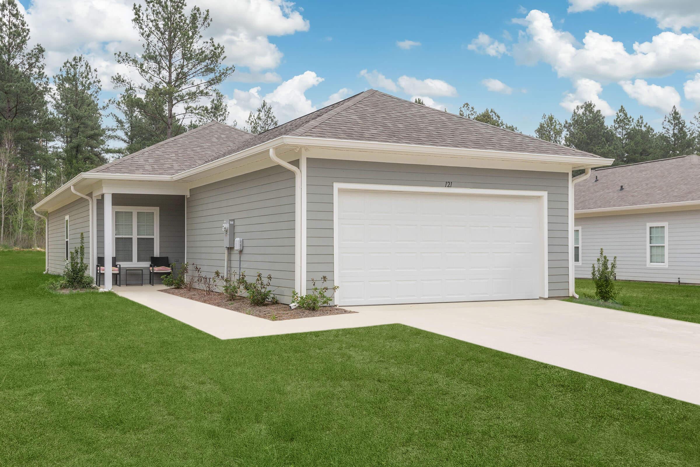 A modern single-story house with light gray siding, a two-car garage, and a neatly manicured lawn. The home features a small front porch with chairs, surrounded by green grass and trees in the background. The sky is blue with fluffy clouds, creating a serene suburban atmosphere.