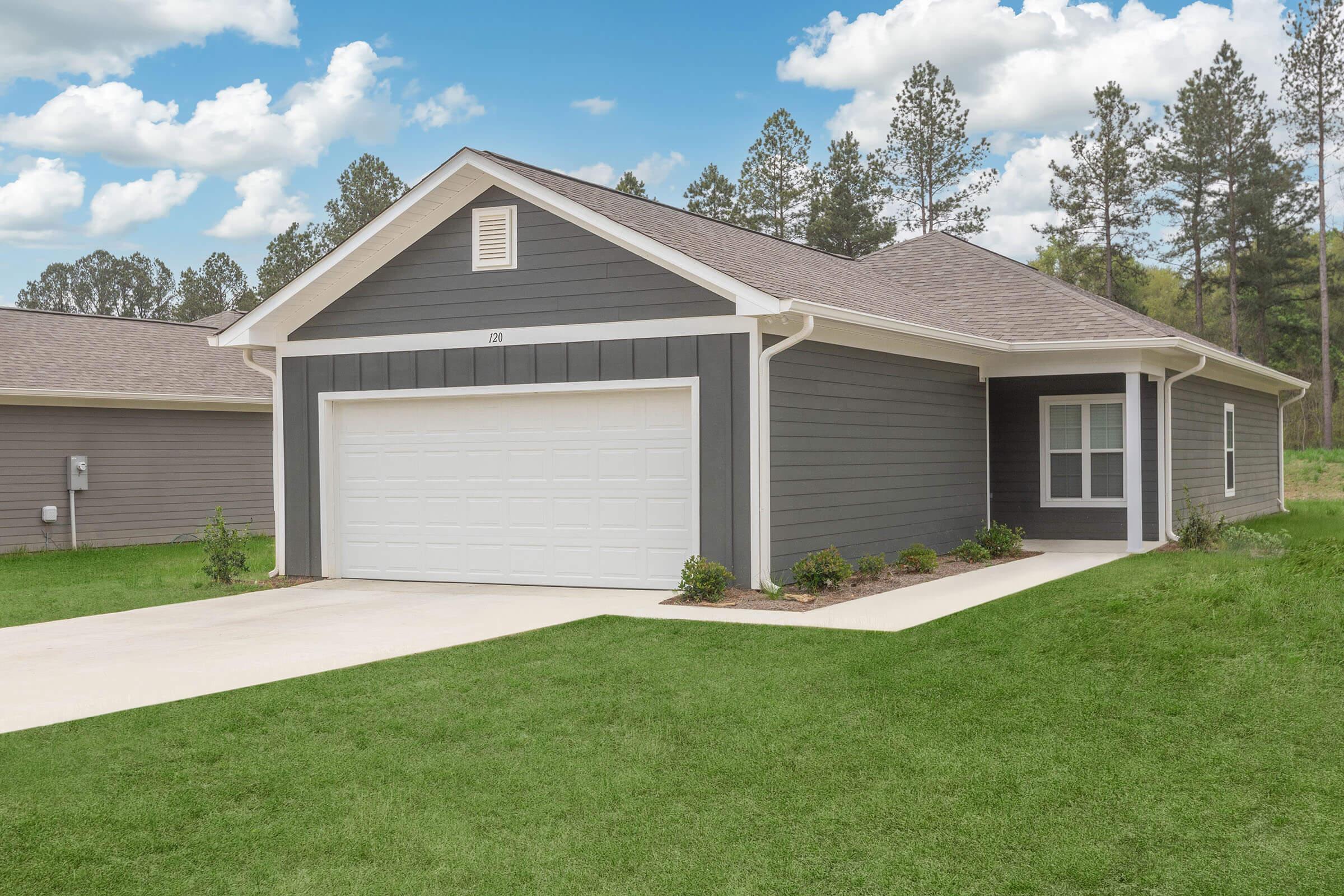 A modern single-story home with gray siding and a white garage door. The front yard is well-maintained with green grass, minimal landscaping, and trees in the background under a partly cloudy blue sky. The house features a porch and large windows.