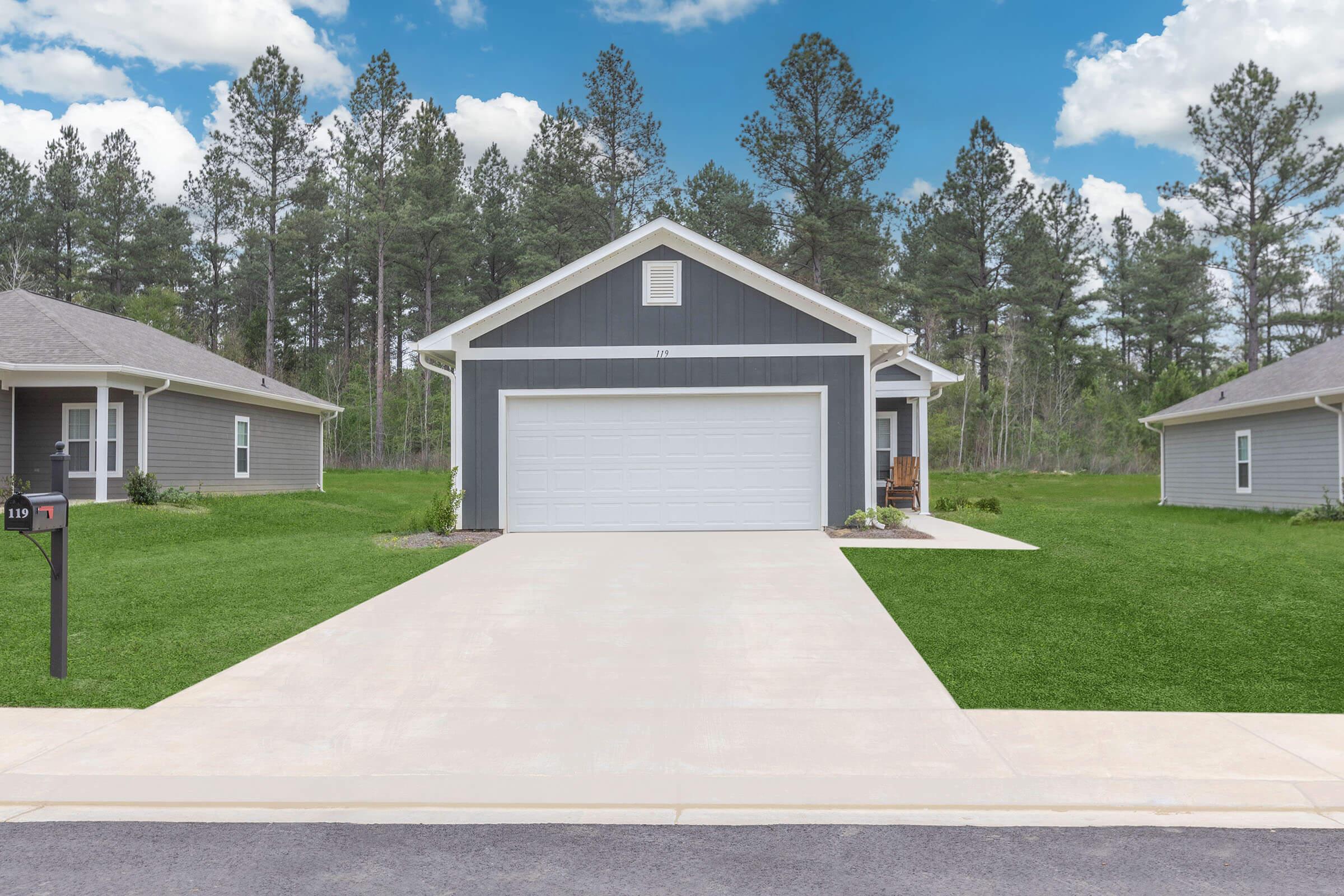 A modern single-story house with gray siding and a white garage door. The driveway is paved, leading to the garage. Lush green grass surrounds the house, with tall trees in the background under a bright blue sky with white clouds. Two similar houses are visible on either side.