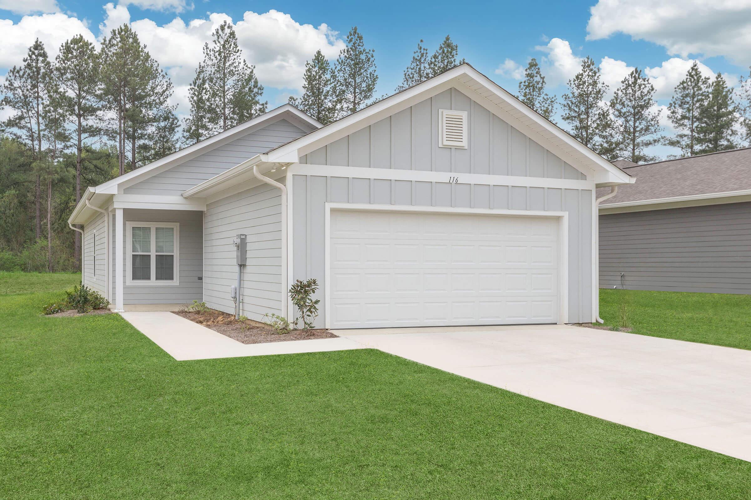 A single-story gray house with a white garage door, situated on a well-maintained lawn. The home has a front porch and a driveway, surrounded by lush green grass and tall pine trees in the background against a clear blue sky.