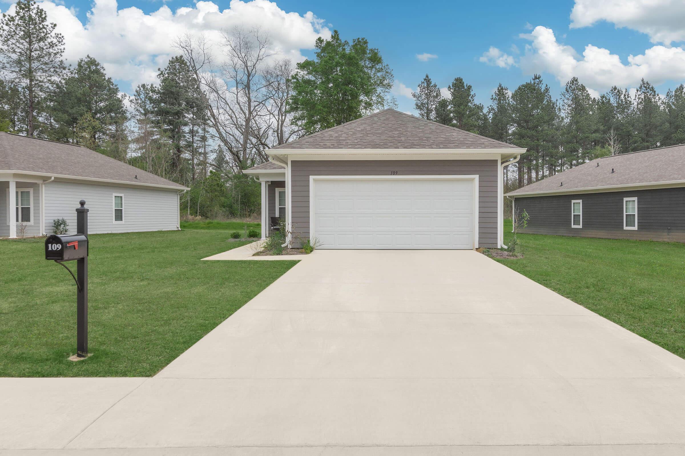 A modern single-family home with a light-colored exterior, a two-car garage, and a concrete driveway. Lush green grass surrounds the house, and there are trees in the background under a partly cloudy sky. A mailbox is visible at the end of the driveway, displaying the number 109.