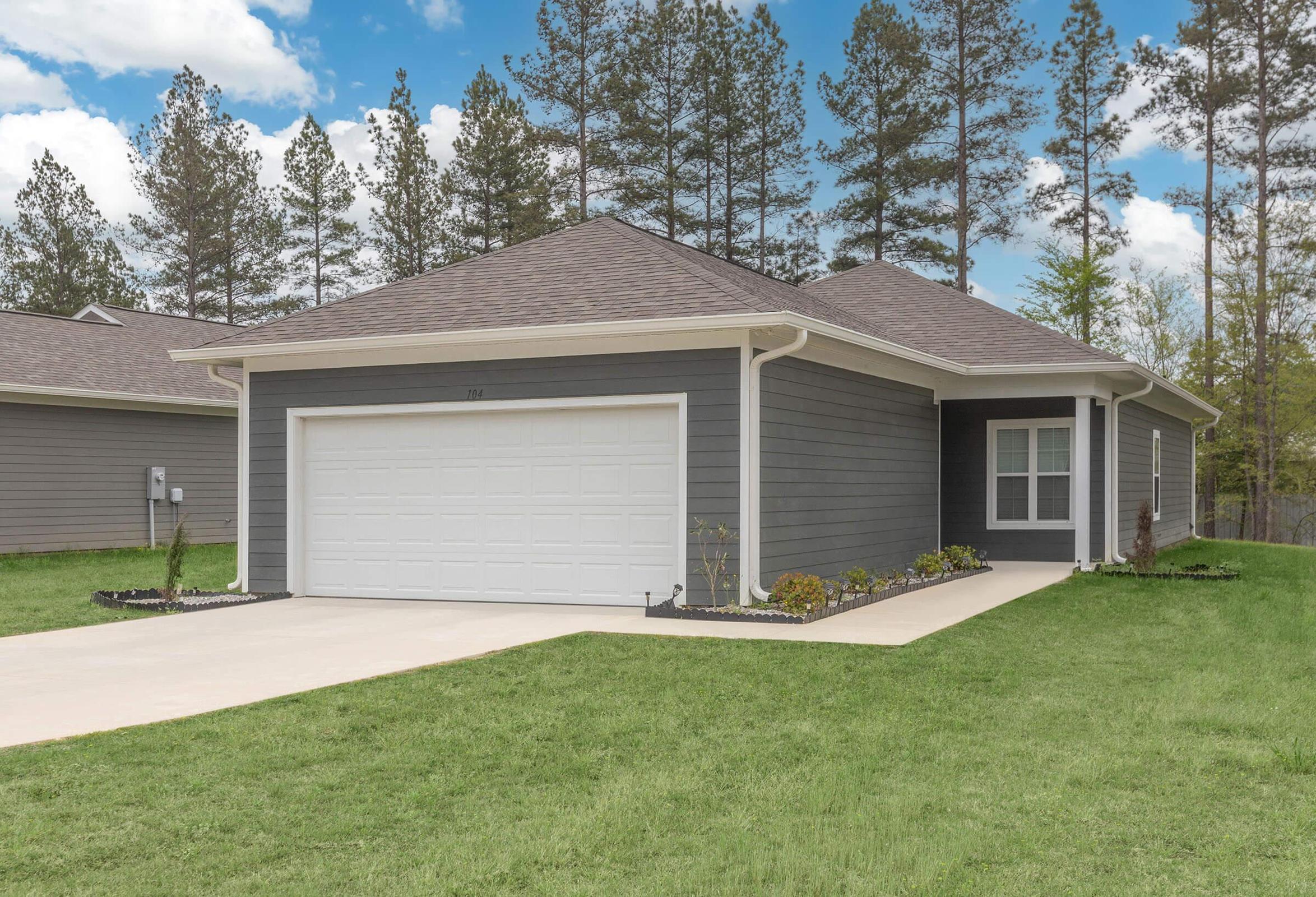 A modern single-story house with gray siding, a sloped roof, and a two-car garage. The lawn is well-maintained with flower beds along the pathway. Tall pine trees are in the background under a partly cloudy sky. The neighborhood has a clean, suburban look.