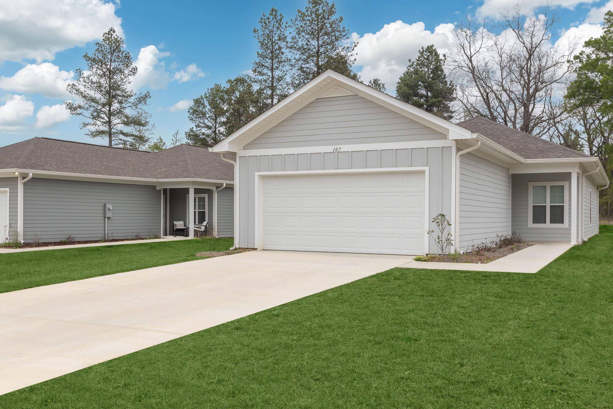 A modern single-story gray house with a well-maintained lawn, featuring a garage with a white door. The background shows a clear blue sky with fluffy clouds and trees. The driveway is paved and leads up to the house, which has a cozy, welcoming appearance.
