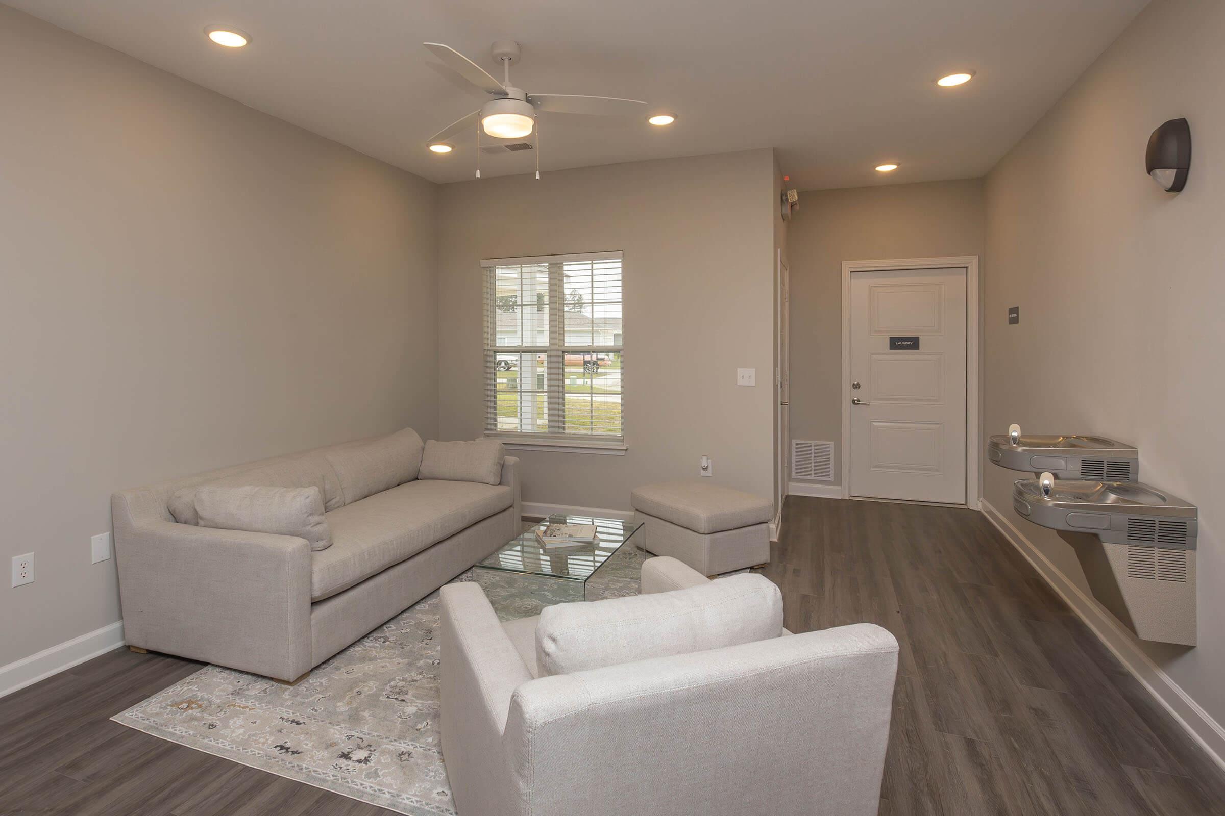 A modern living room featuring a light gray sofa set, matching ottoman, and a glass coffee table on a decorative rug. Large windows allow natural light, and the room has a ceiling fan for ventilation. A solid door is present, along with a water fountain, adding convenience to the space.