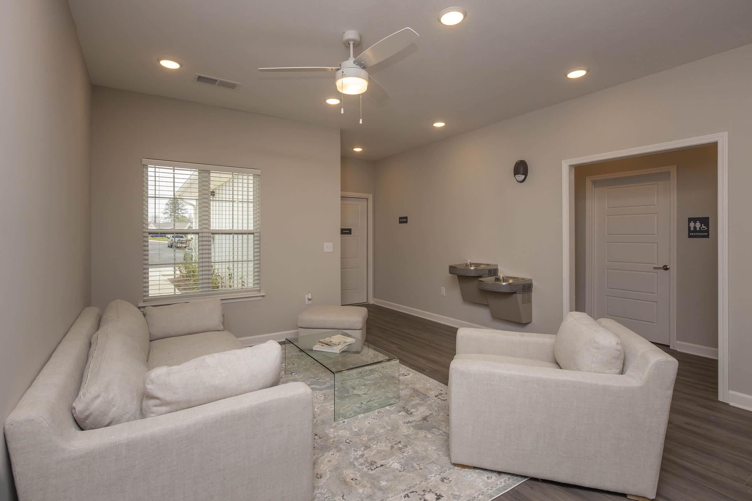 A modern, minimalist living space featuring a light-colored sofa set arranged around a glass coffee table, with a rug underneath. The room has gray walls, a ceiling fan, and large windows allowing natural light. A water fountain is visible on the wall, and a door leads to a restroom.