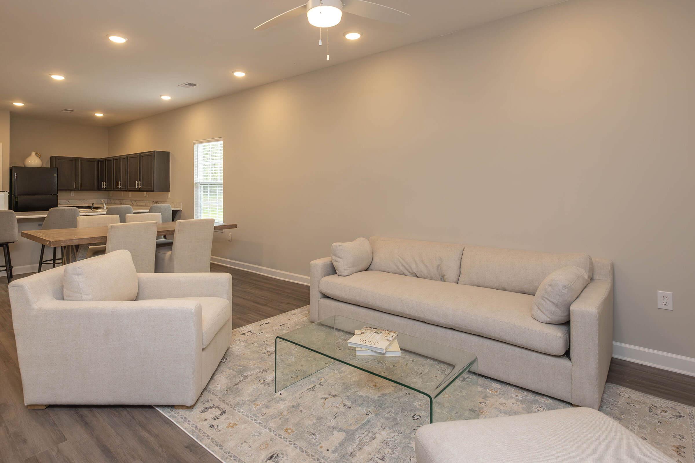 A modern living room featuring a light-colored sofa and two armchairs, complemented by a glass coffee table on a patterned area rug. The walls are painted a soft gray, and there are bright lights illuminating the space. In the background, a dining area with a table and chairs can be seen.