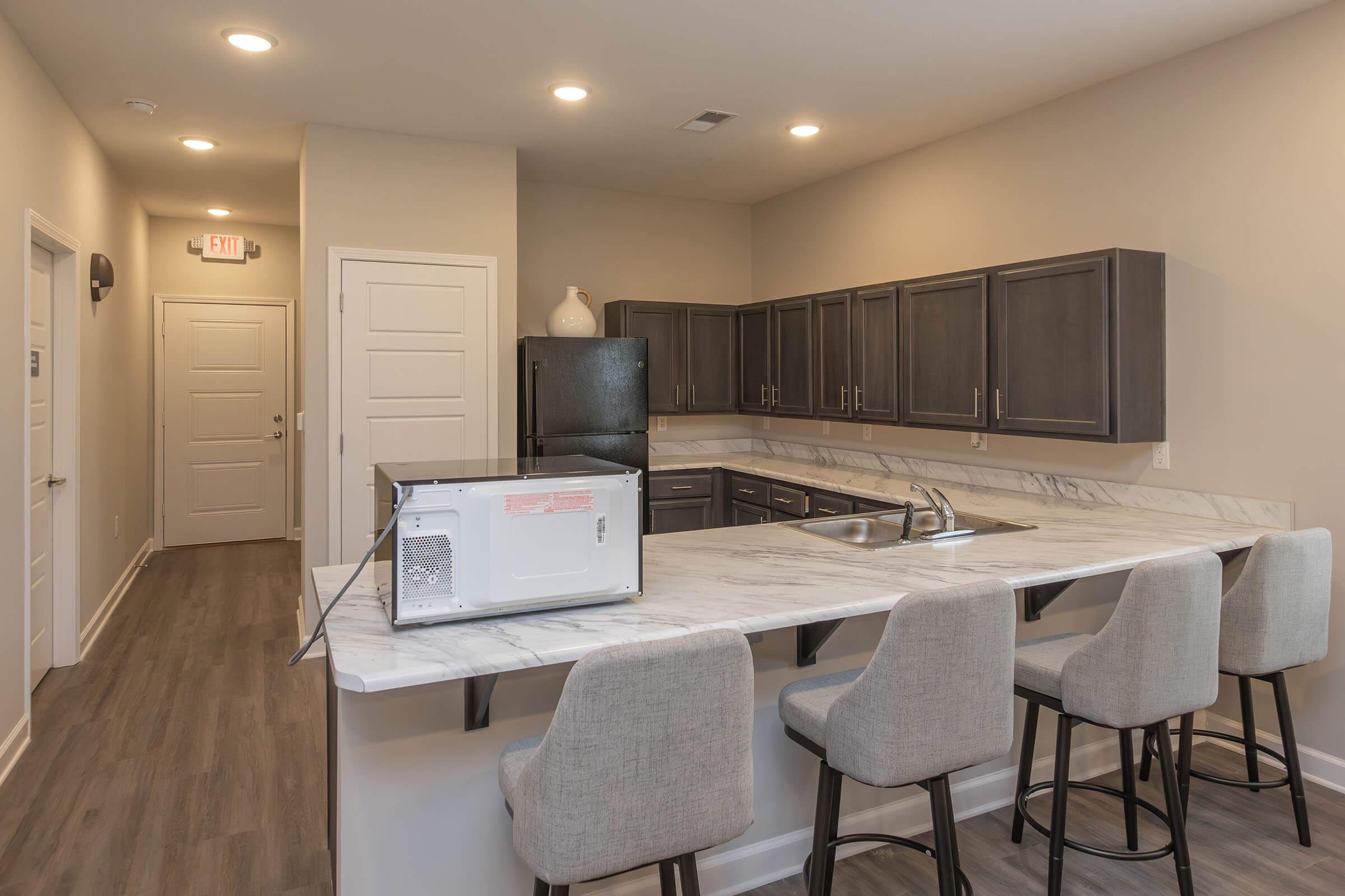 Modern kitchen with dark cabinets, light marble countertop, and bar seating for four. Features a black refrigerator and a microwave on the countertop. The space is well-lit with recessed lighting and has a neutral color scheme. An entry door is visible in the background.