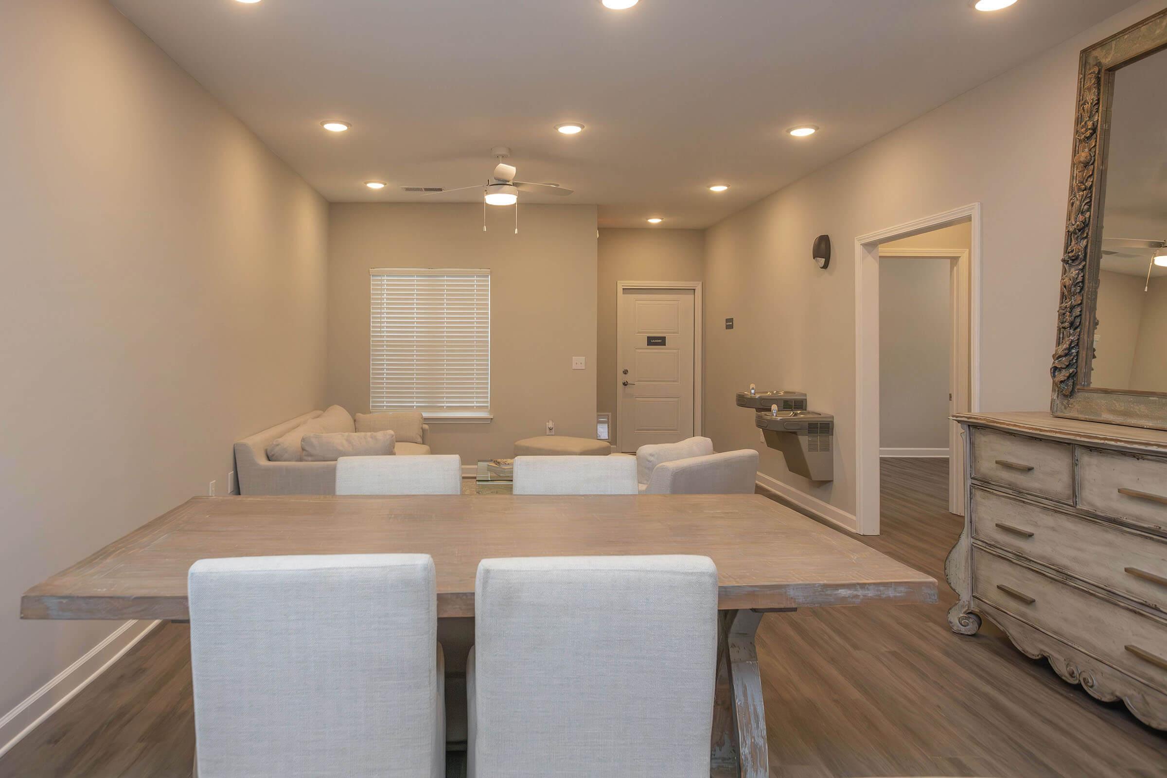 A modern, minimalist interior featuring a dining table with upholstered chairs, a light-colored couch, and a decorative mirror on the wall. The room has neutral-colored walls, a ceiling fan, and ample natural light from a window, with a small bathroom or utility area visible in the background.
