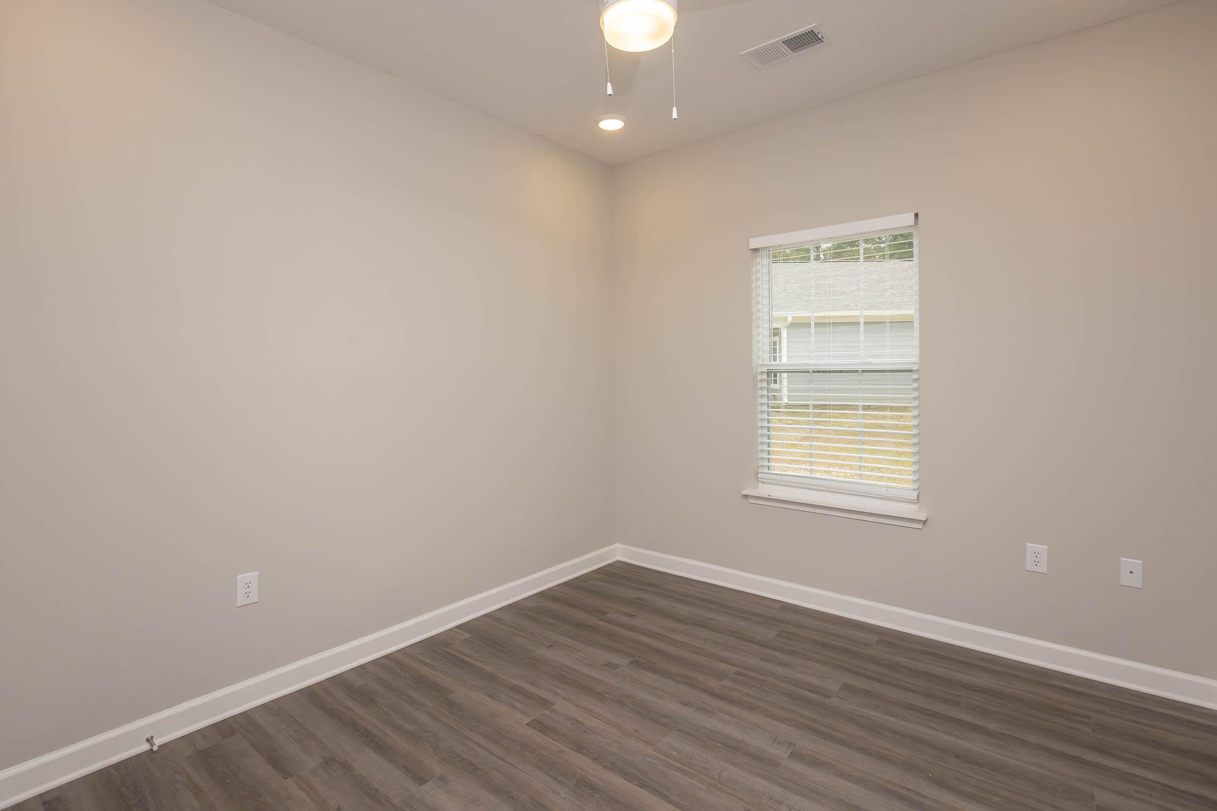 A minimalistic room with light gray walls, a ceiling fan, and a window with blinds. The floor is covered in wood-like laminate, and the area is well-lit with natural light coming through the window. The space is empty, suggesting it could be used for various purposes.