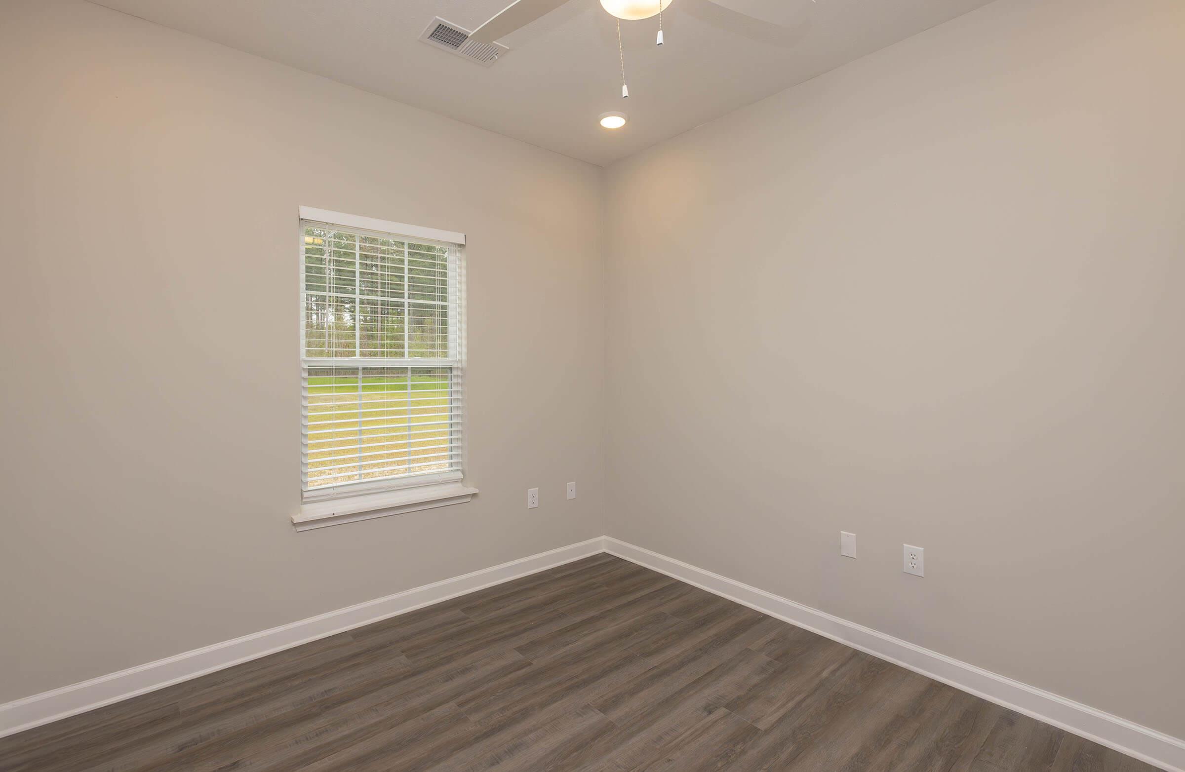 A small, empty room with light gray walls and a wooden floor. There is a single window with white blinds, allowing natural light to enter. The ceiling features a ceiling fan, and the room is devoid of furniture, giving it a clean and minimalist appearance.