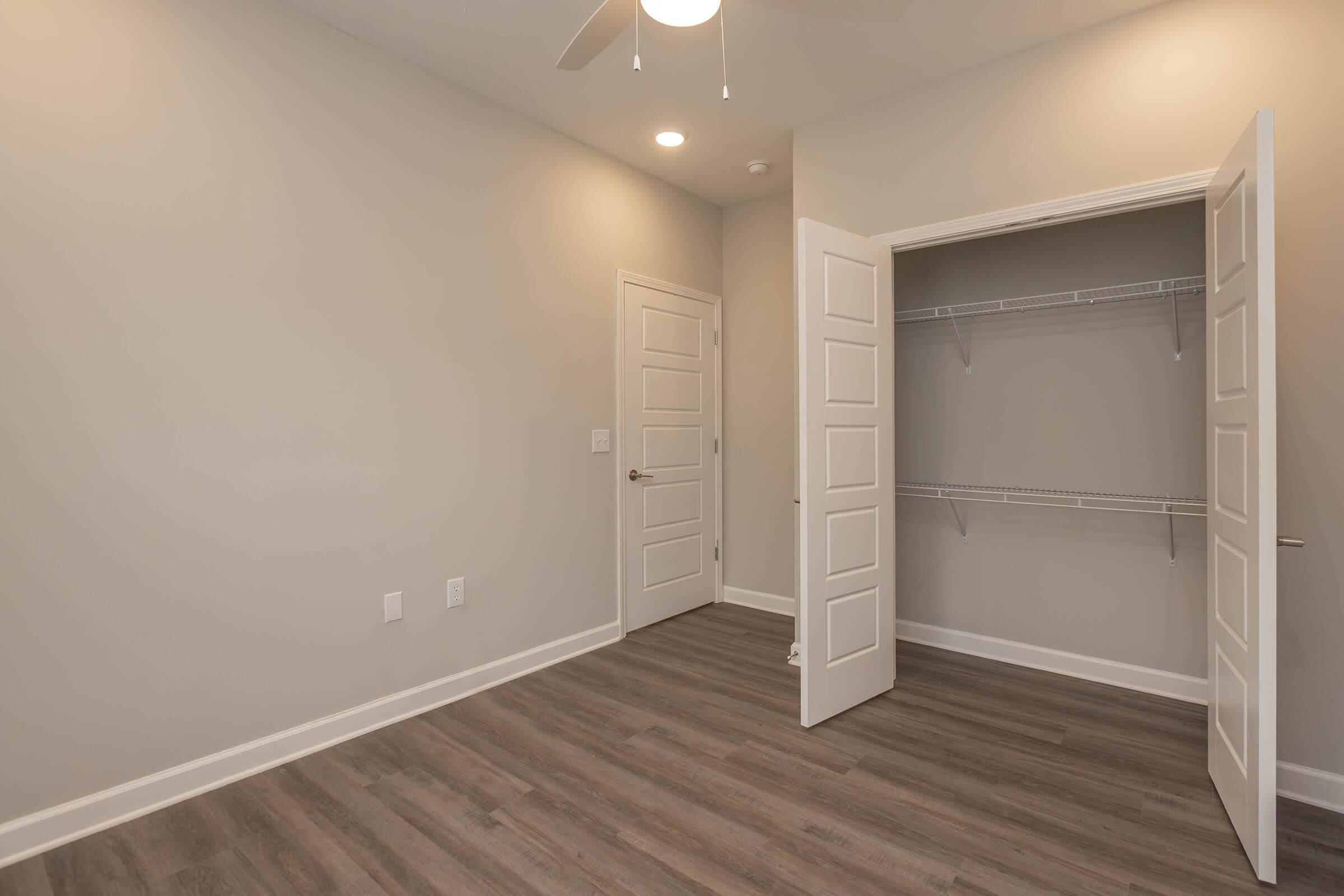 A spacious, empty room with light gray walls and a ceiling fan. There's a double door closet on one side, featuring wire shelving. The flooring is a warm wood tone, and the room is well-lit by ceiling lights. The overall ambiance is clean and modern, suitable for various uses.