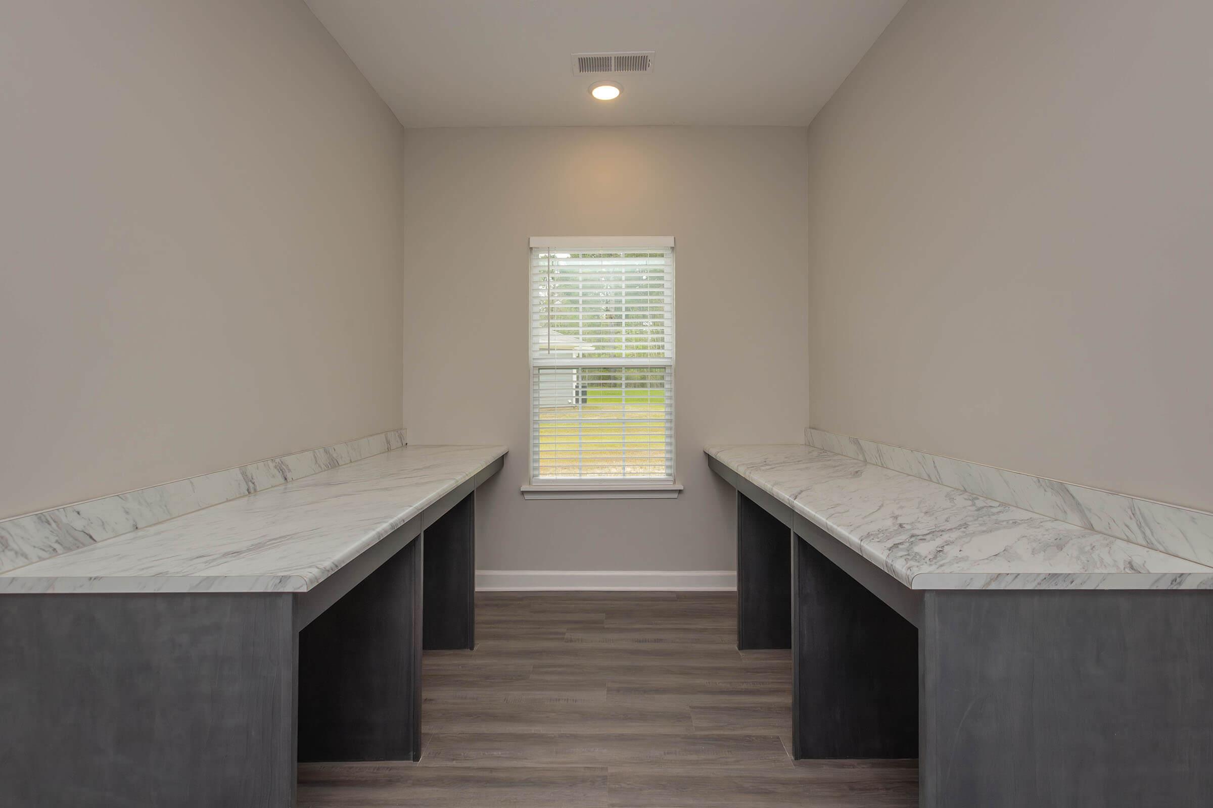 A bright, minimalist room featuring two marble-topped counters with dark cabinetry below. There is a large window with blinds allowing natural light to enter, illuminating the space. The walls are painted a light color, and the flooring is a modern wood-like material.