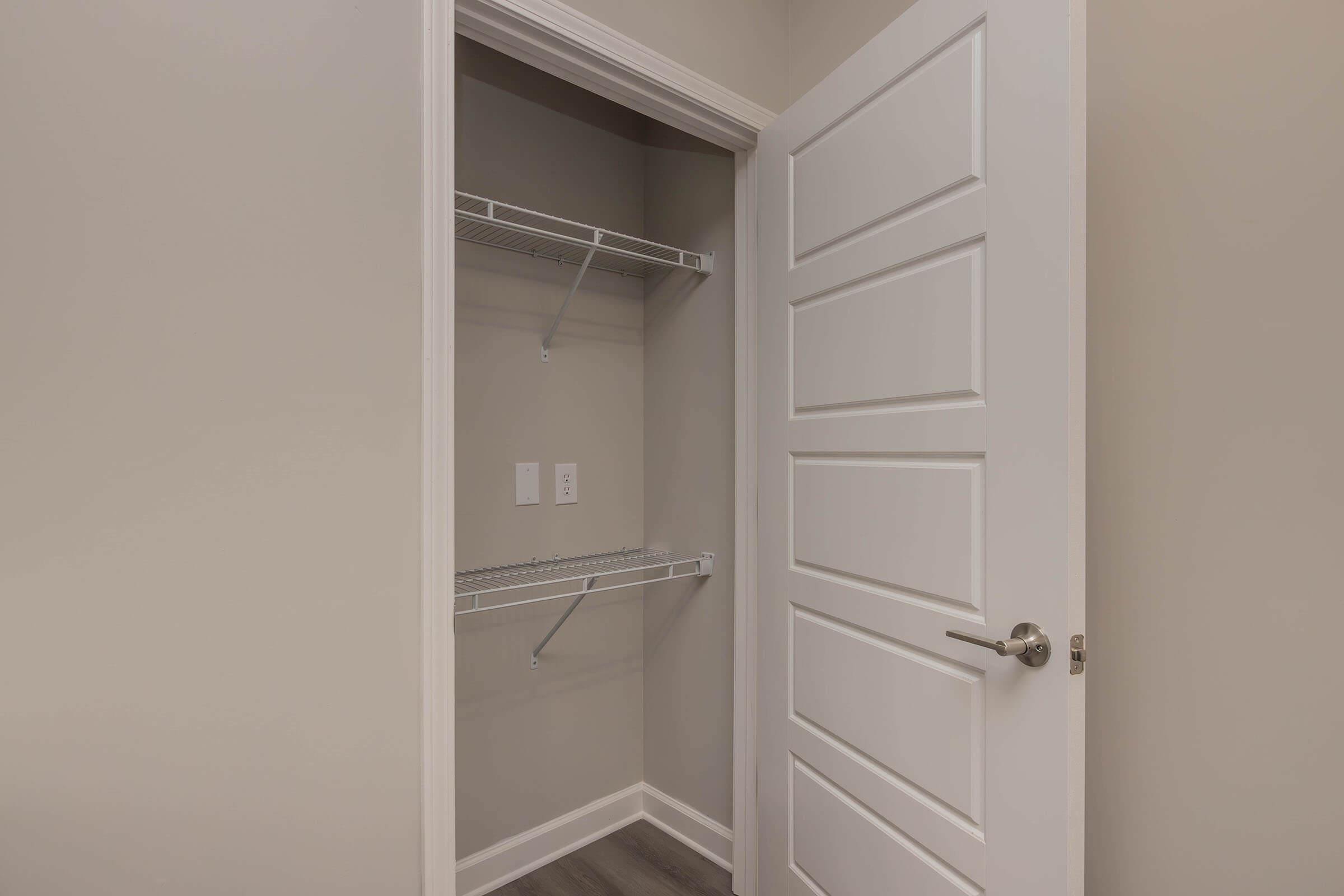 A closet with empty metal shelving and a closed white door. The walls are painted a light color, and the floor is covered with gray laminate. Electrical outlets are visible on the wall inside the closet. The door has a modern design with horizontal panels.