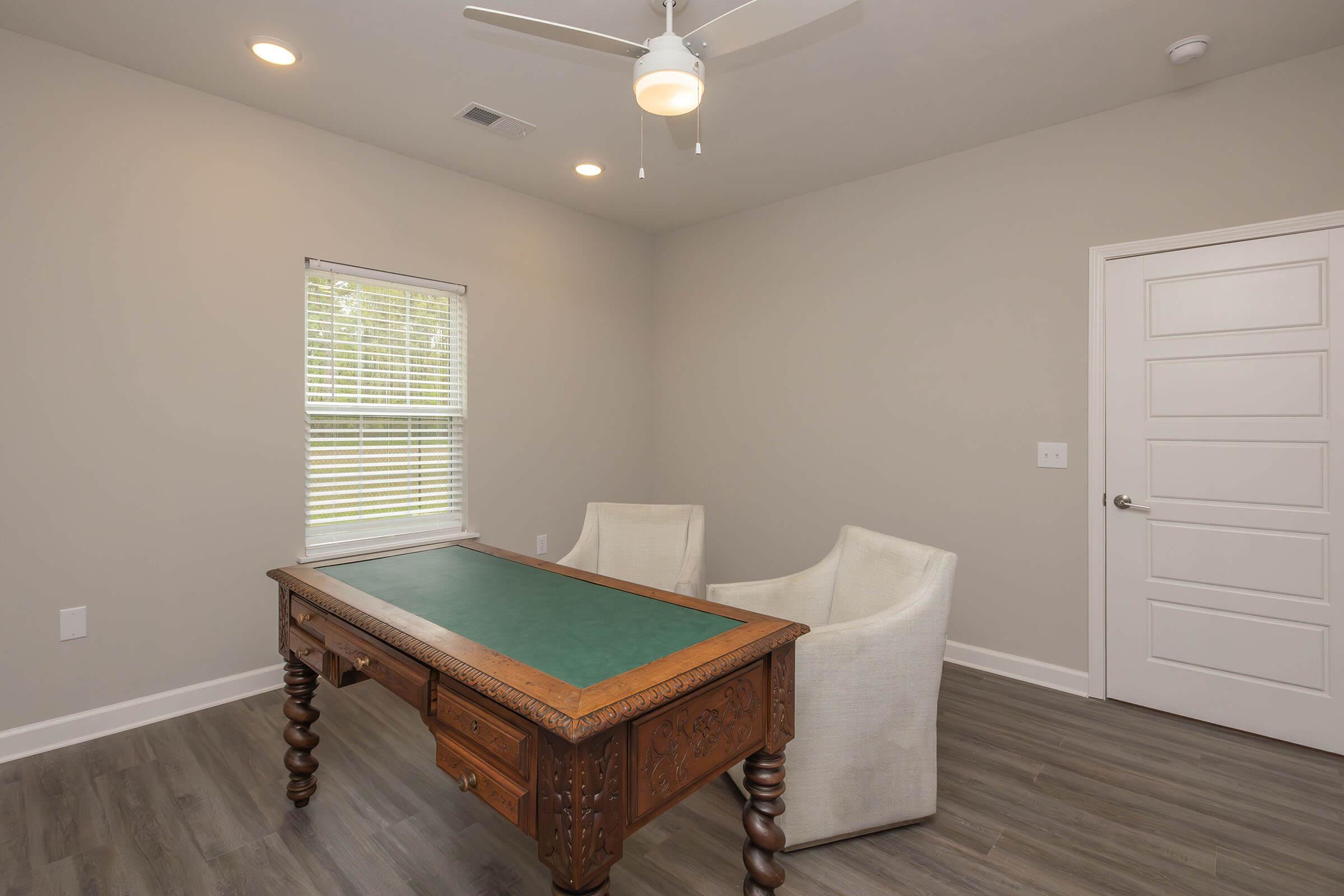 A well-lit home office featuring a vintage wooden desk with a green tabletop, two white upholstered chairs, a ceiling fan, and a window with blinds. The walls are painted in a neutral gray tone, and the flooring is a light wood finish.