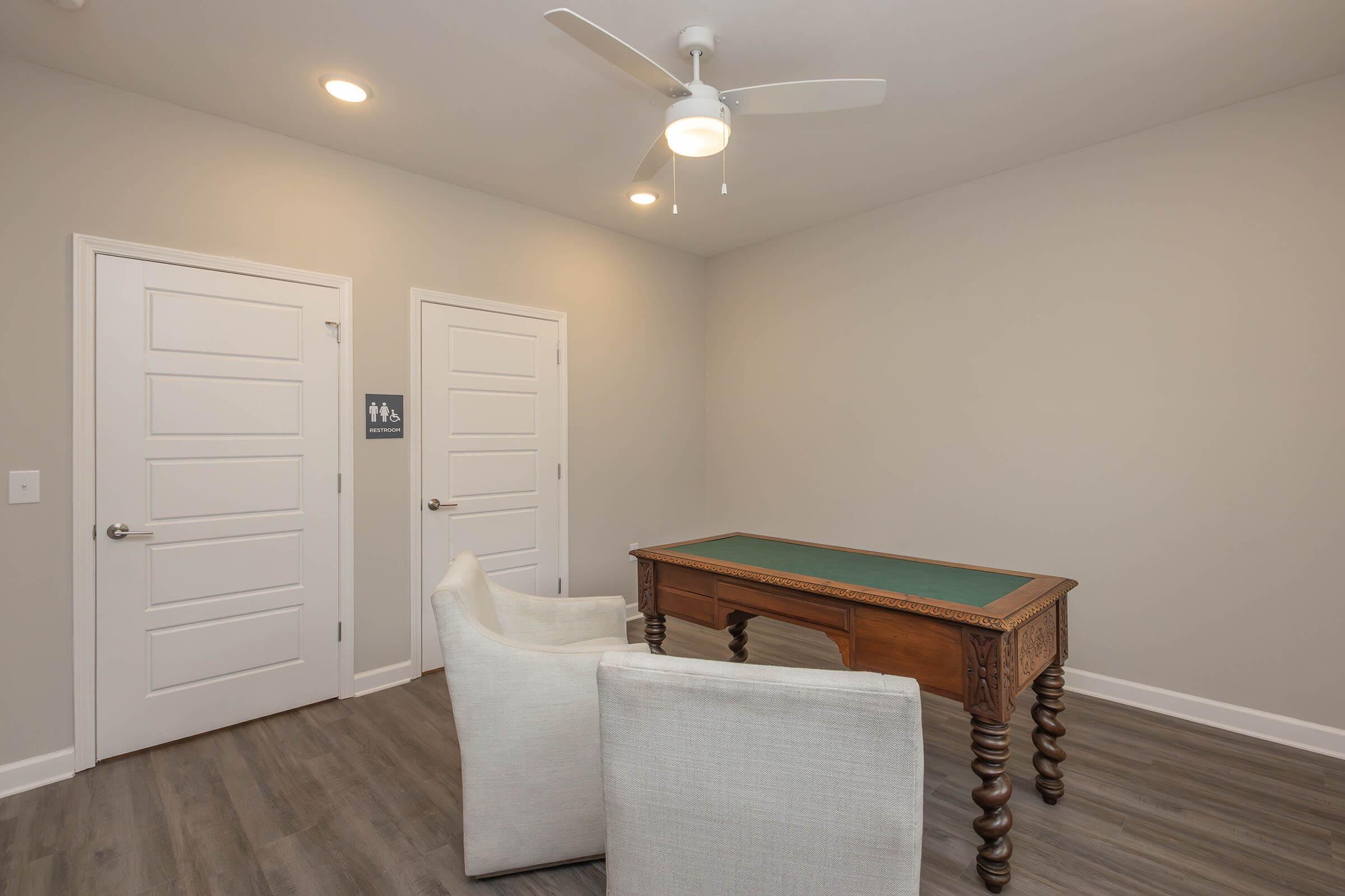 A simple, well-lit room featuring a wooden table with a green tabletop, two white upholstered chairs, and two doors along the wall. A ceiling fan is installed above, and there are signs indicating restrooms nearby. The walls are painted in a neutral color, giving a clean and modern appearance.