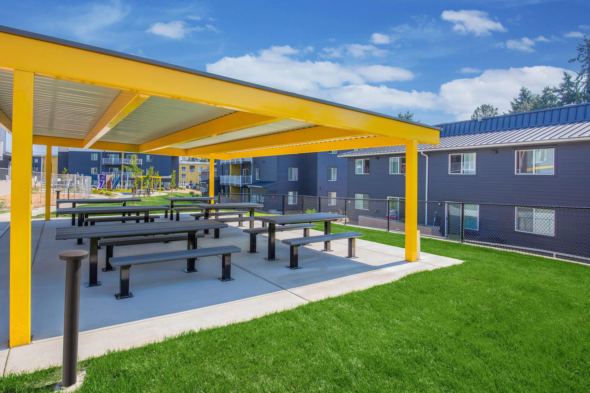 A bright yellow covered area with picnic-style tables, surrounded by green grass. In the background, there are modern apartment buildings with blue and gray exteriors. The scene is set under a clear blue sky, creating a cheerful and inviting outdoor space.
