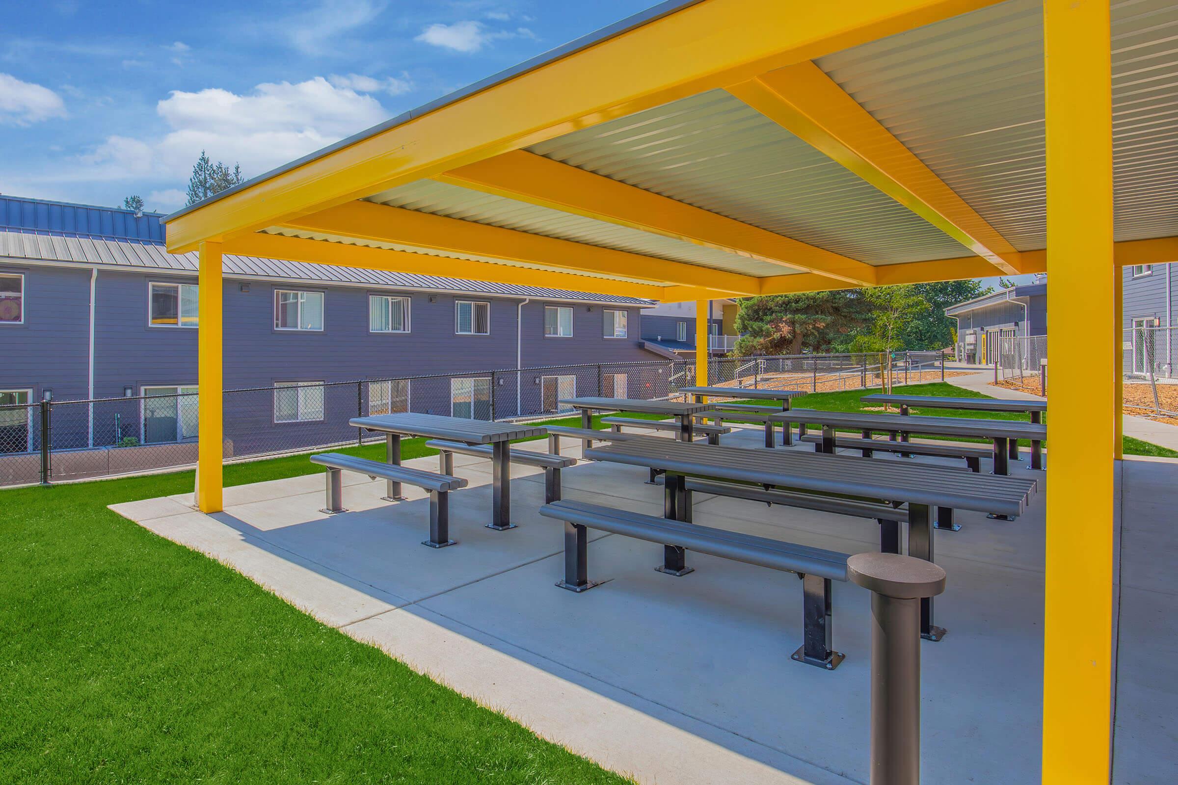 A bright, yellow covered outdoor seating area with multiple picnic-style tables on a concrete slab. The background features gray buildings and a fenced lawn under a clear blue sky, creating a welcoming environment for gatherings or events.