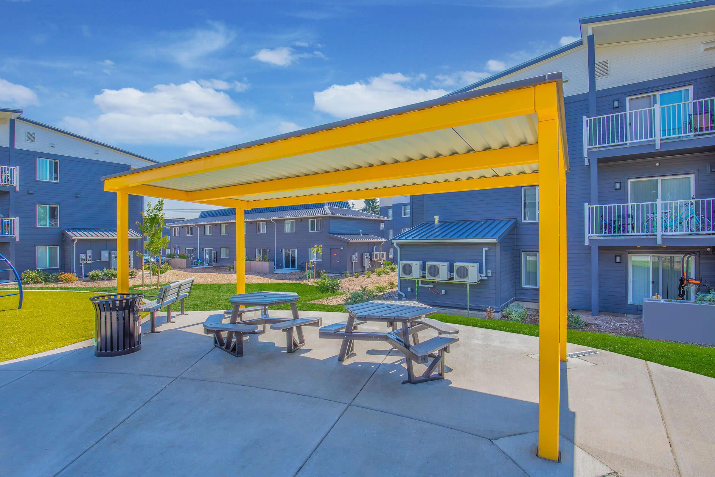 A vibrant yellow pergola providing shade over picnic tables in a landscaped area. Surrounding the space are modern apartment buildings with balconies and air conditioning units. The sky is clear with a few clouds, creating a bright and inviting atmosphere.