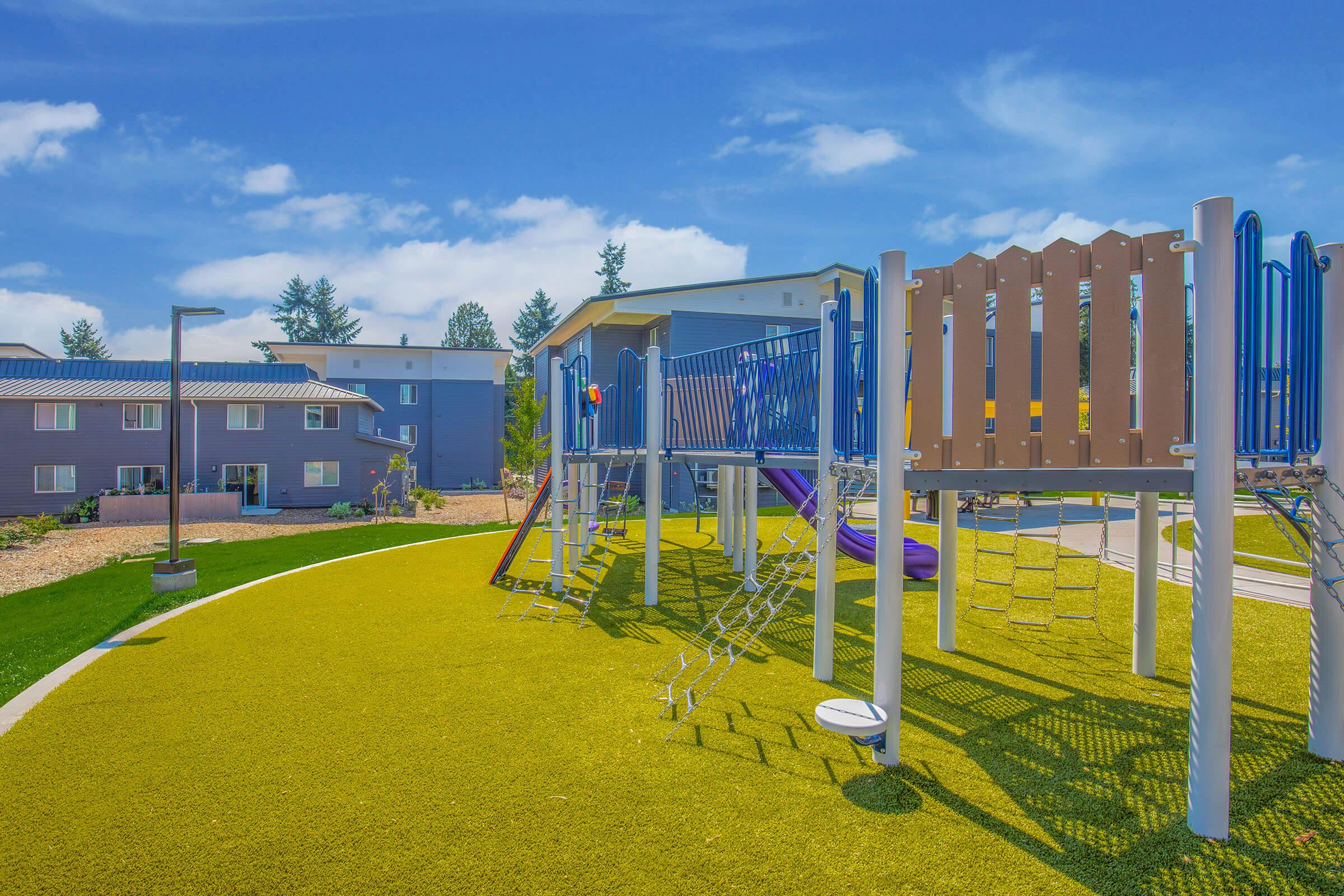 A colorful playground with slides, climbing structures, and green turf surrounded by modern buildings. The sky is bright blue with a few clouds, creating a cheerful atmosphere for outdoor play.
