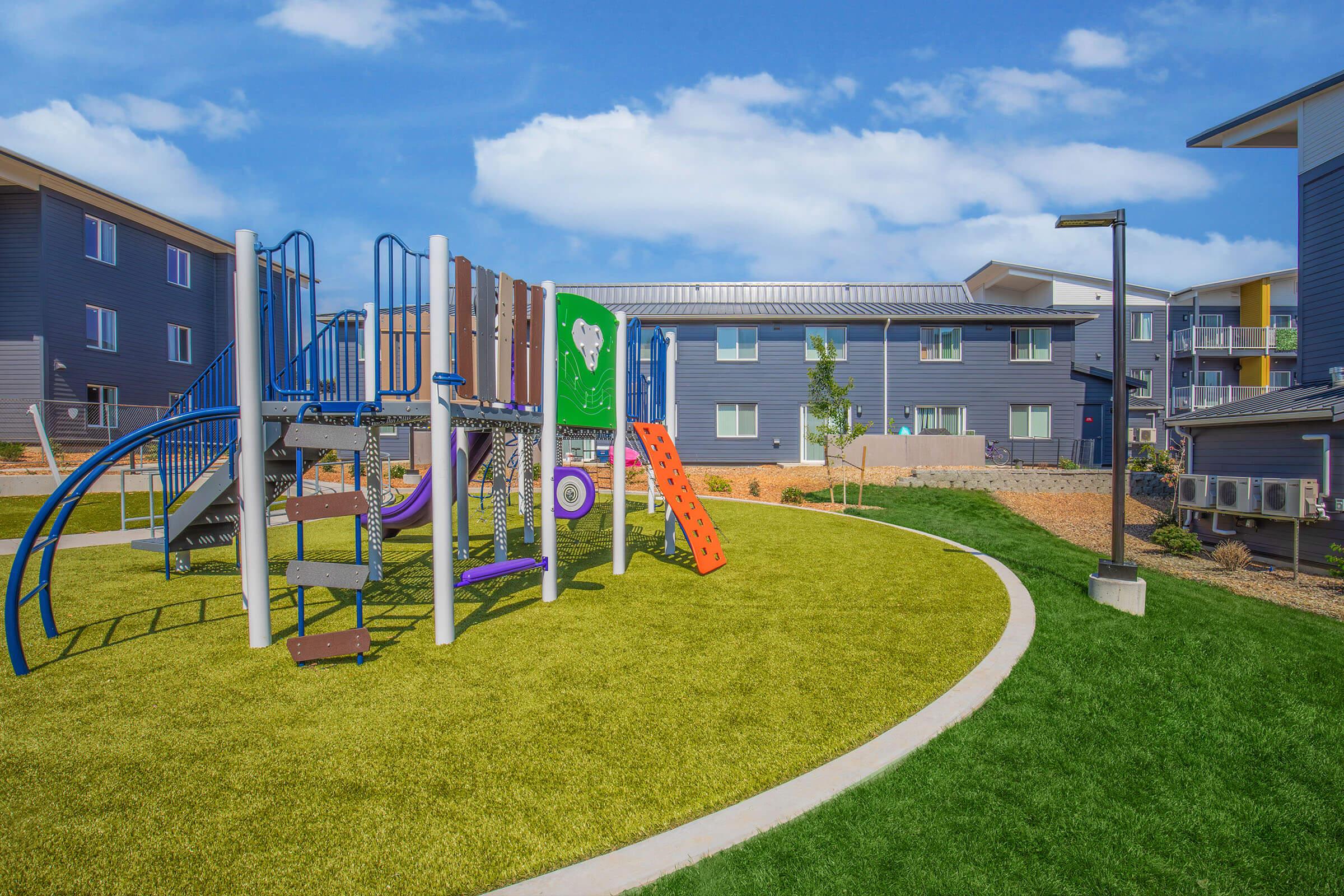 Colorful playground equipment, including slides and climbing structures, on artificial grass surrounded by modern apartment buildings. The sky is bright with a few clouds, creating a cheerful outdoor environment.